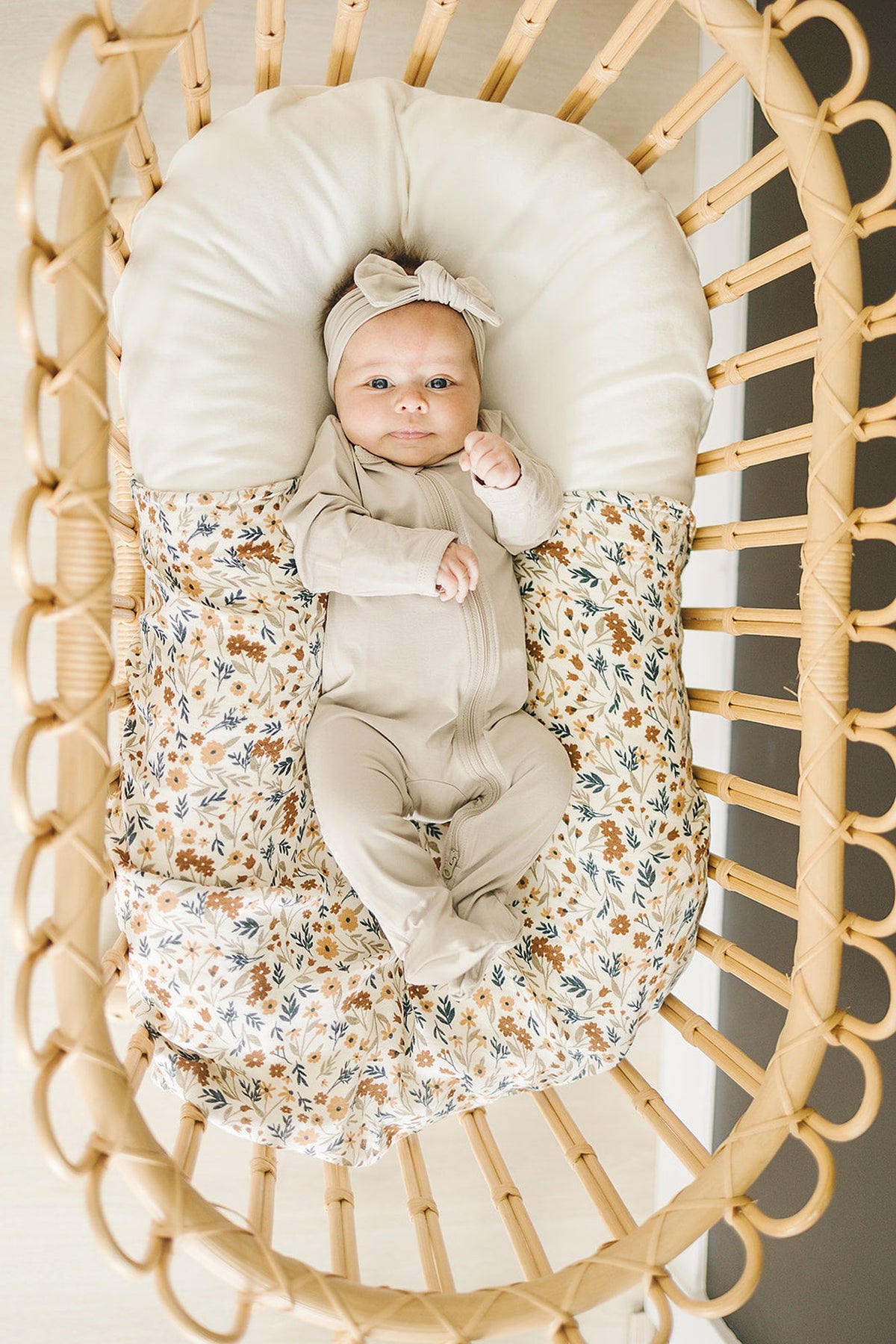 A baby in a beige footed zipper pajama lies in a floral-patterned bassinet, wearing a matching headband.