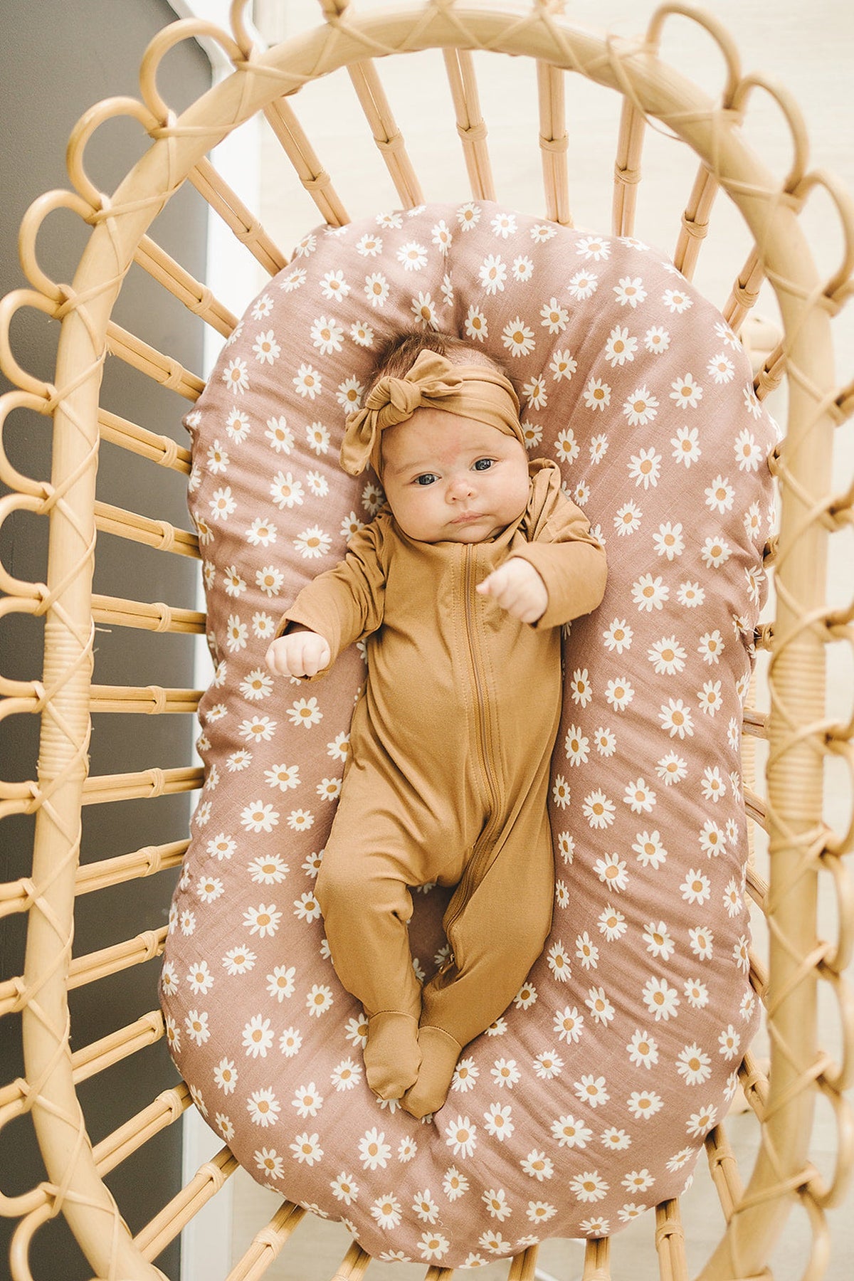 Baby in a mustard footed pajama with a headband, lying on a floral cushion in a wicker crib.