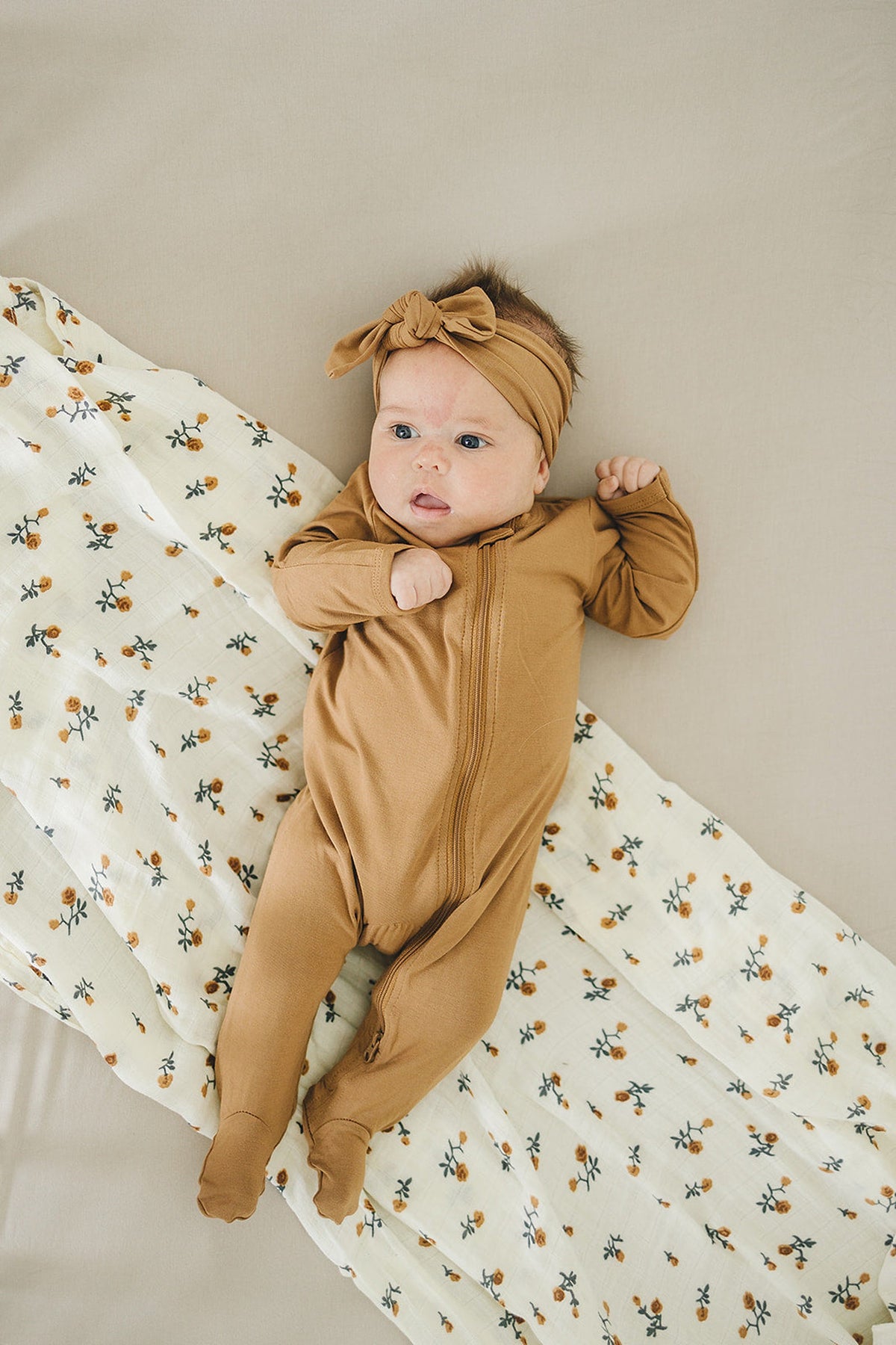 A baby in a mustard footed pajama with a matching headband lies on a floral-patterned blanket.