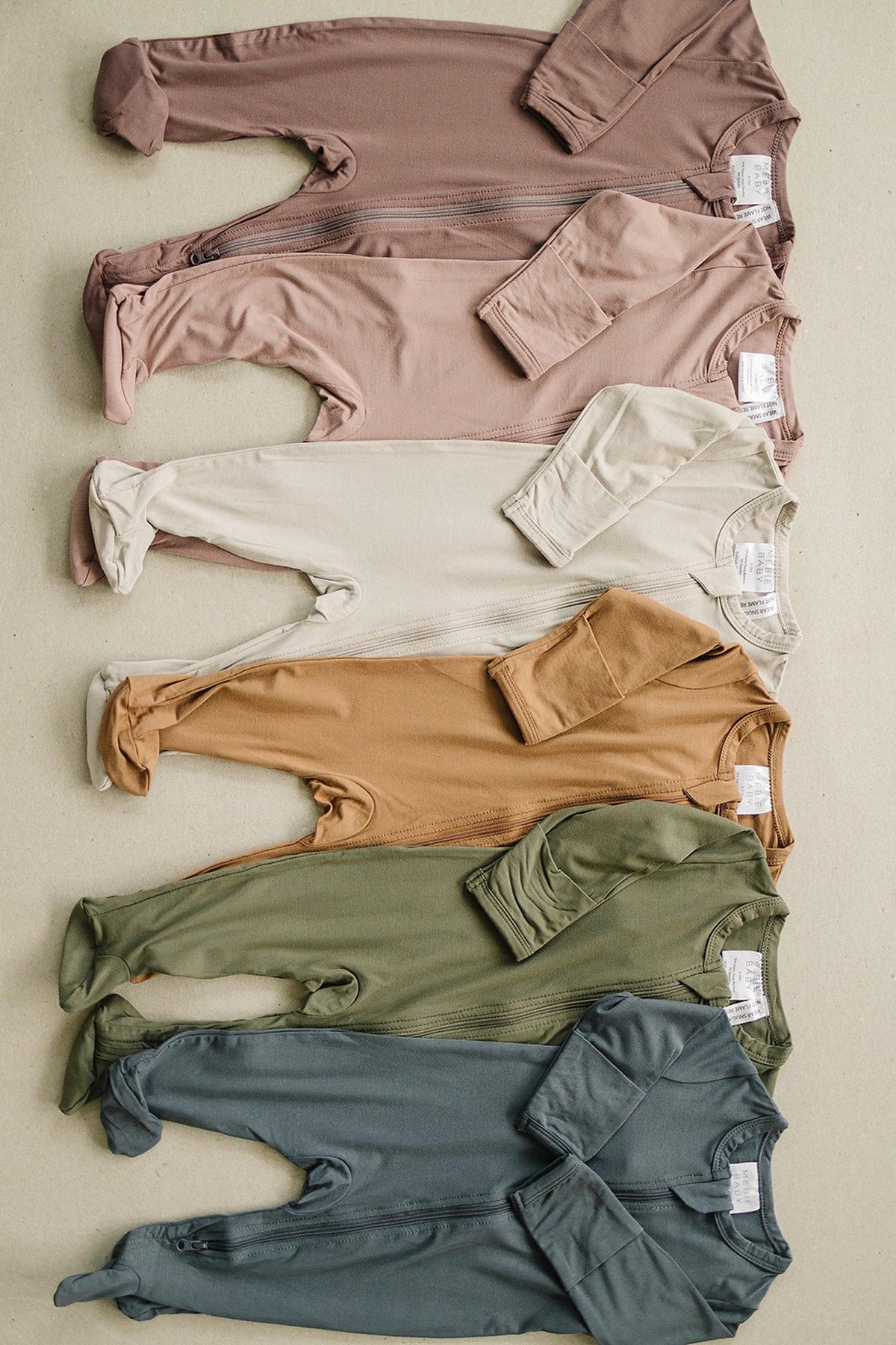 Five bamboo footed zipper pajamas in plum, cream, mustard, olive, and teal colors, laid flat on a surface.