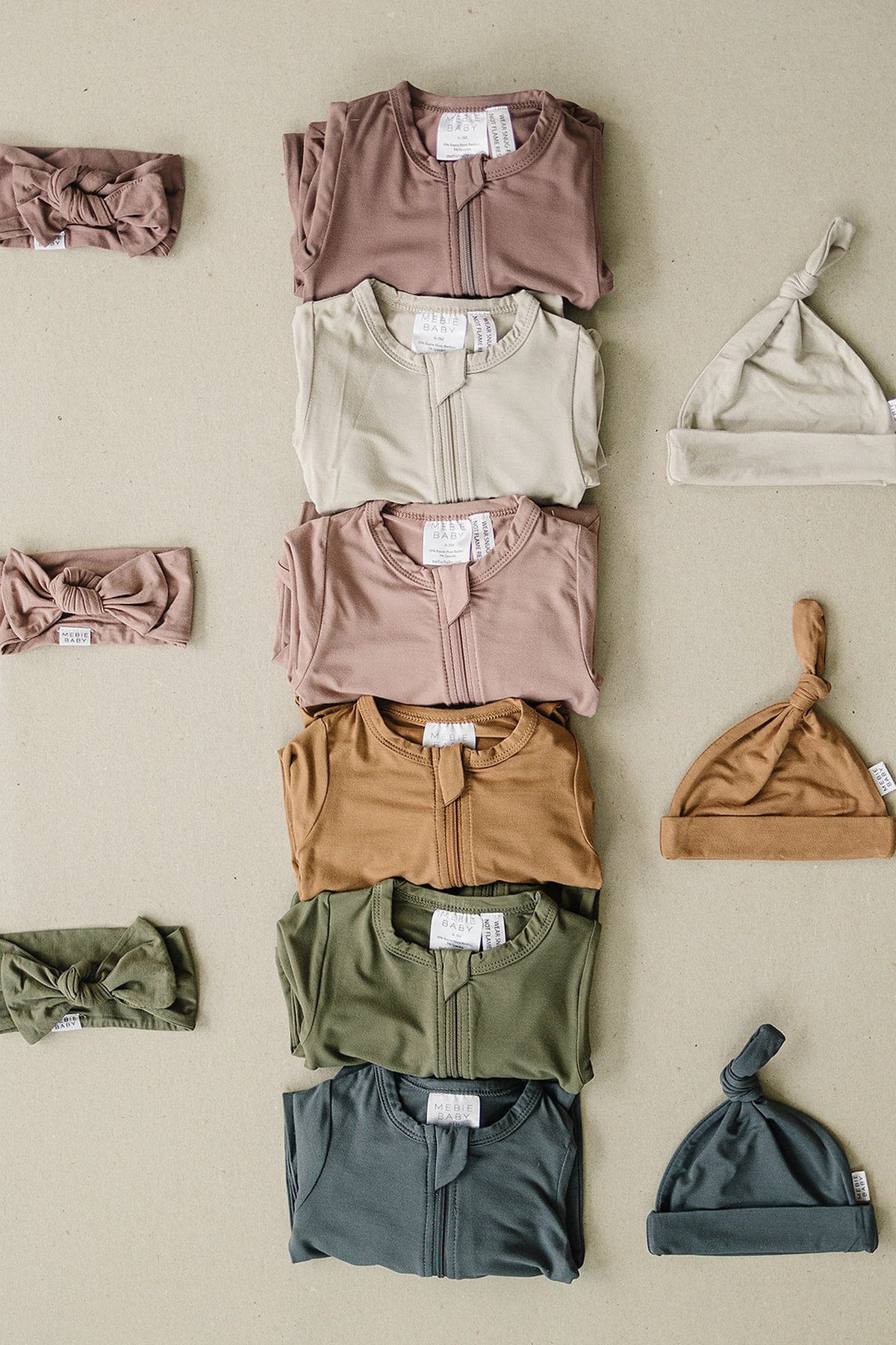 Assorted Mebie Baby bamboo pajamas in neutral tones, with matching hats and headbands, displayed on a beige background.