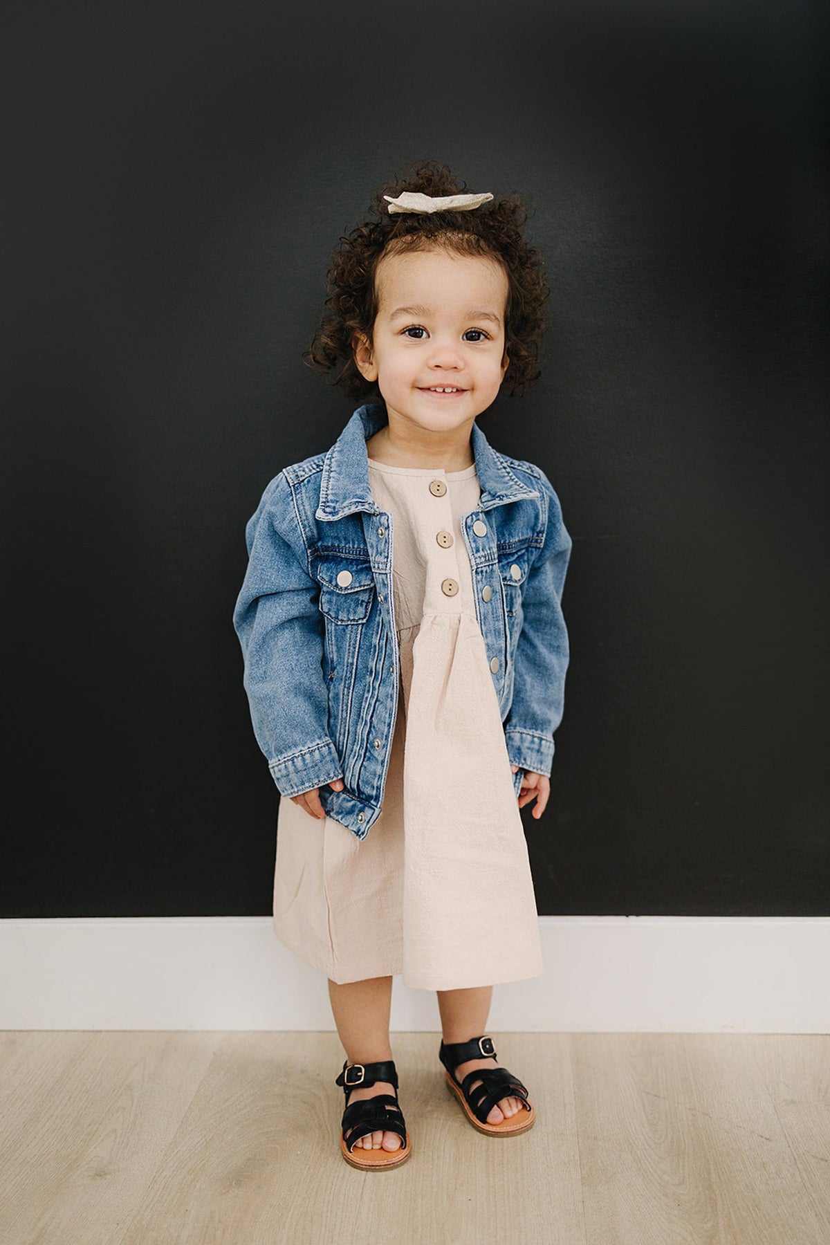 Toddler wearing a blue denim jacket and beige dress, standing against a black wall, with curly hair and sandals.