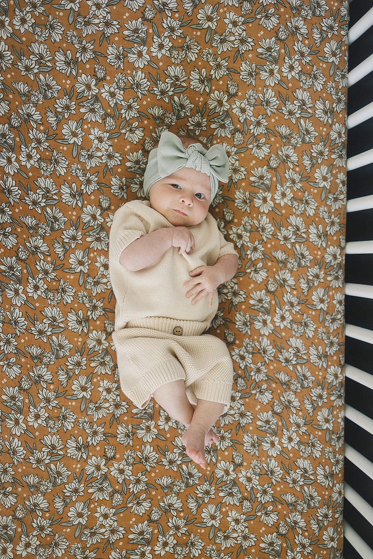 A baby in beige clothing lies on a floral-patterned crib sheet with a magnolia design.