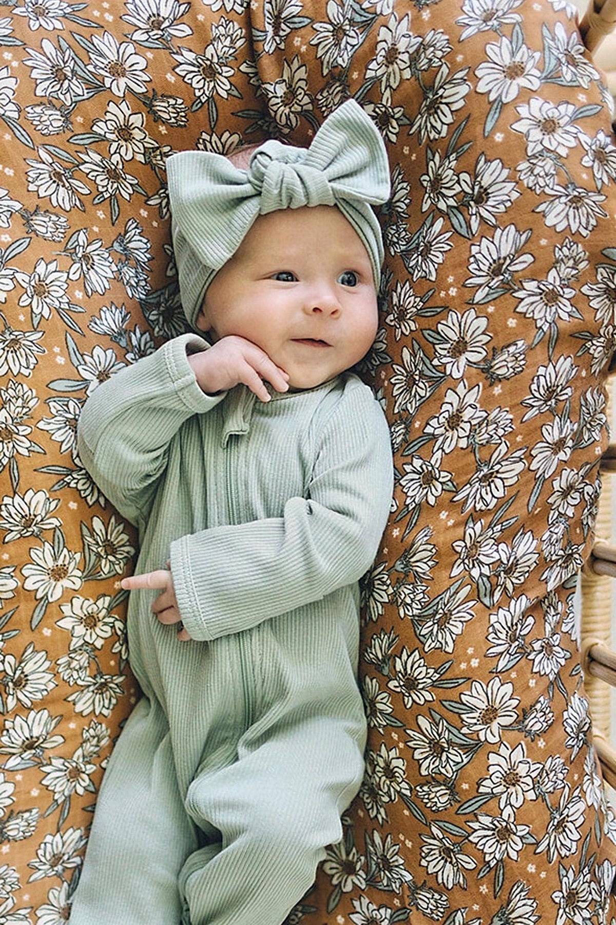 A baby wearing a sage ribbed head wrap and onesie lies on a floral-patterned cushion.