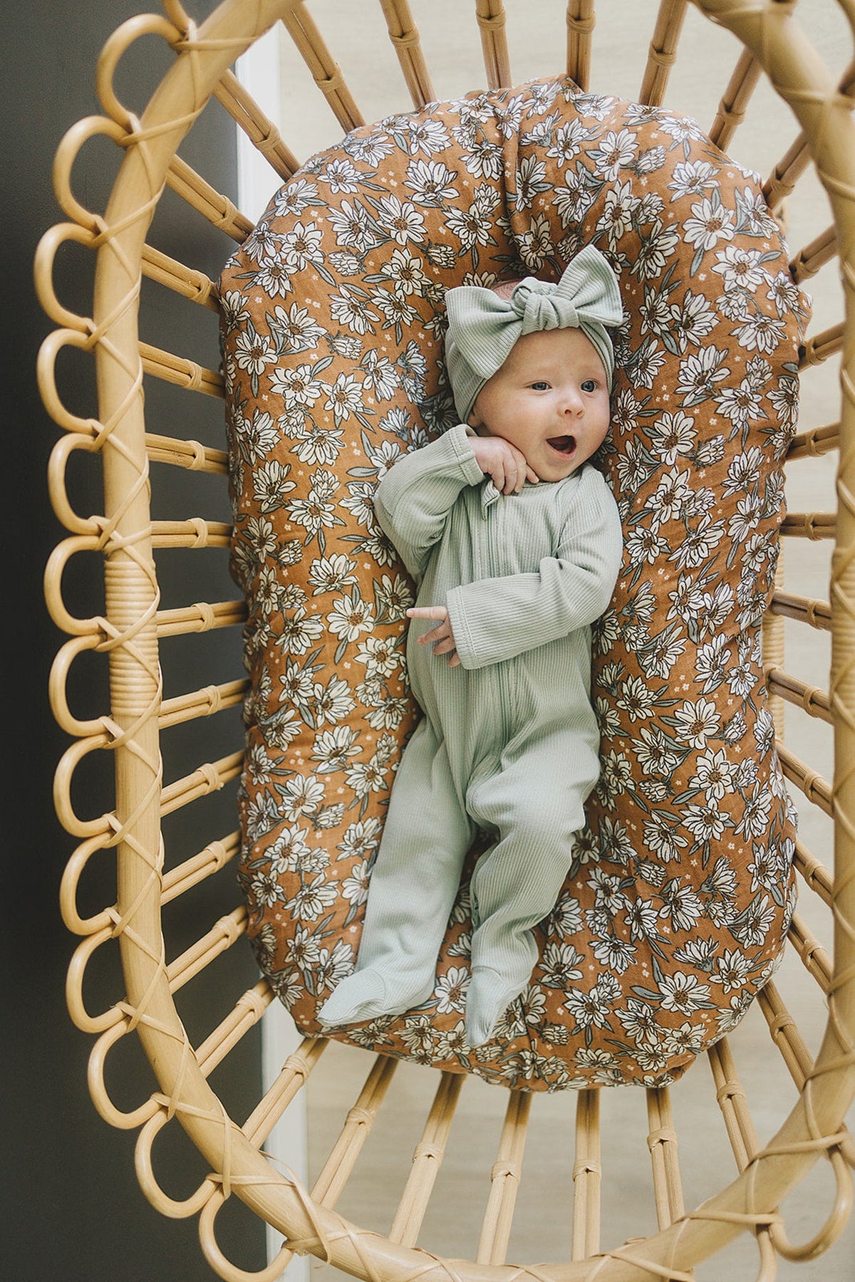 A baby wearing sage ribbed pajamas and headband lies on a floral cushion in a wicker bassinet.