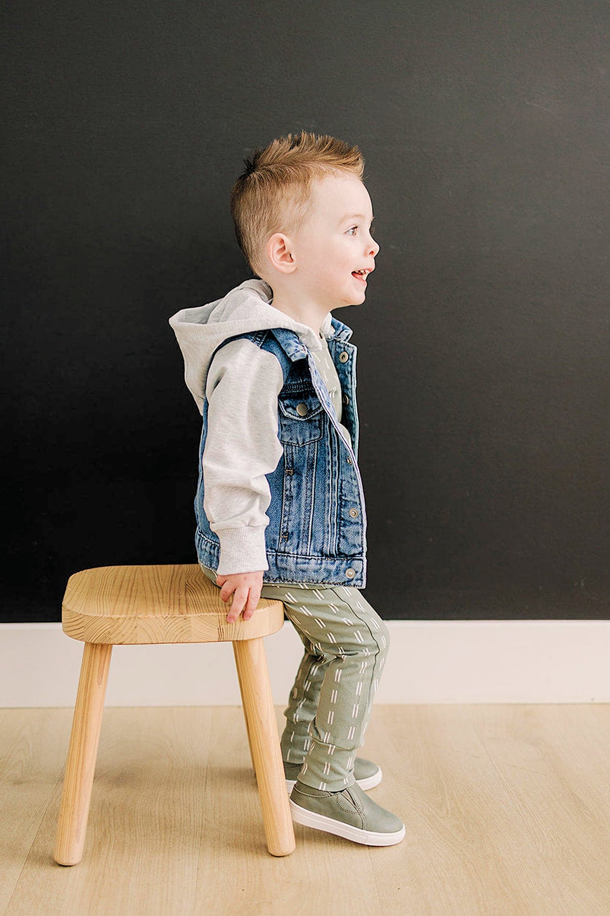 A child wearing a denim jacket with a grey hood sits on a wooden stool, smiling against a dark background.