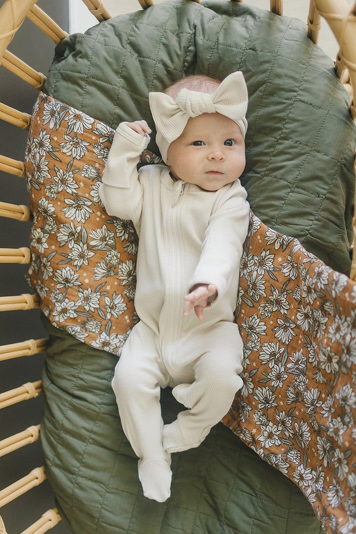 Baby in white outfit and bow headband lies on a green and floral quilt, surrounded by wicker chair.