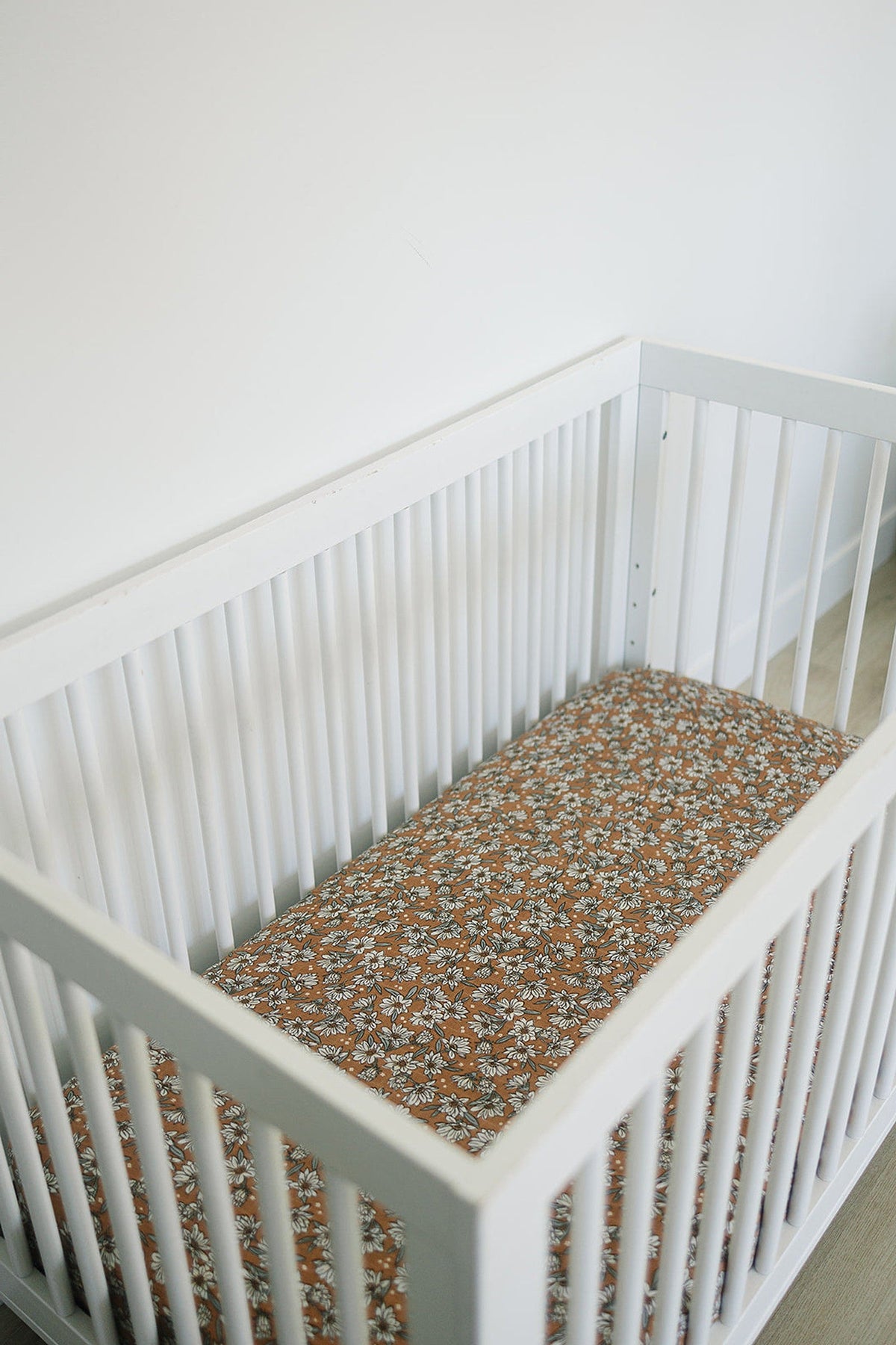 White crib with a floral-patterned muslin crib sheet in warm tones, set in a minimalistic nursery.
