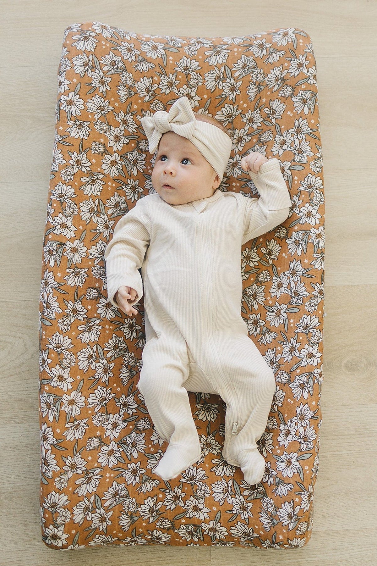 A baby in a cream outfit lies on a floral-patterned changing pad cover with white and brown flowers.