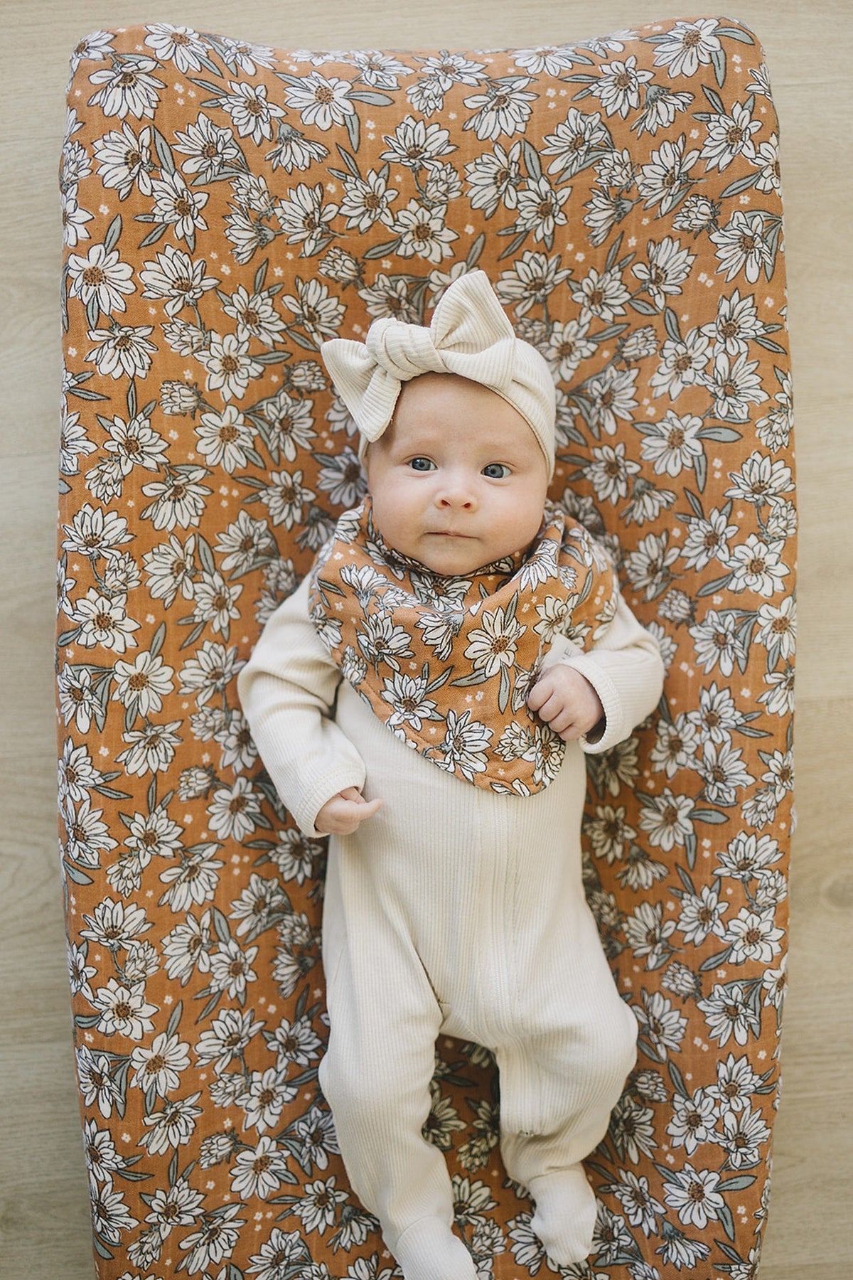 Baby in cream outfit and headband on floral patterned mat, wearing a matching bib with white flowers.