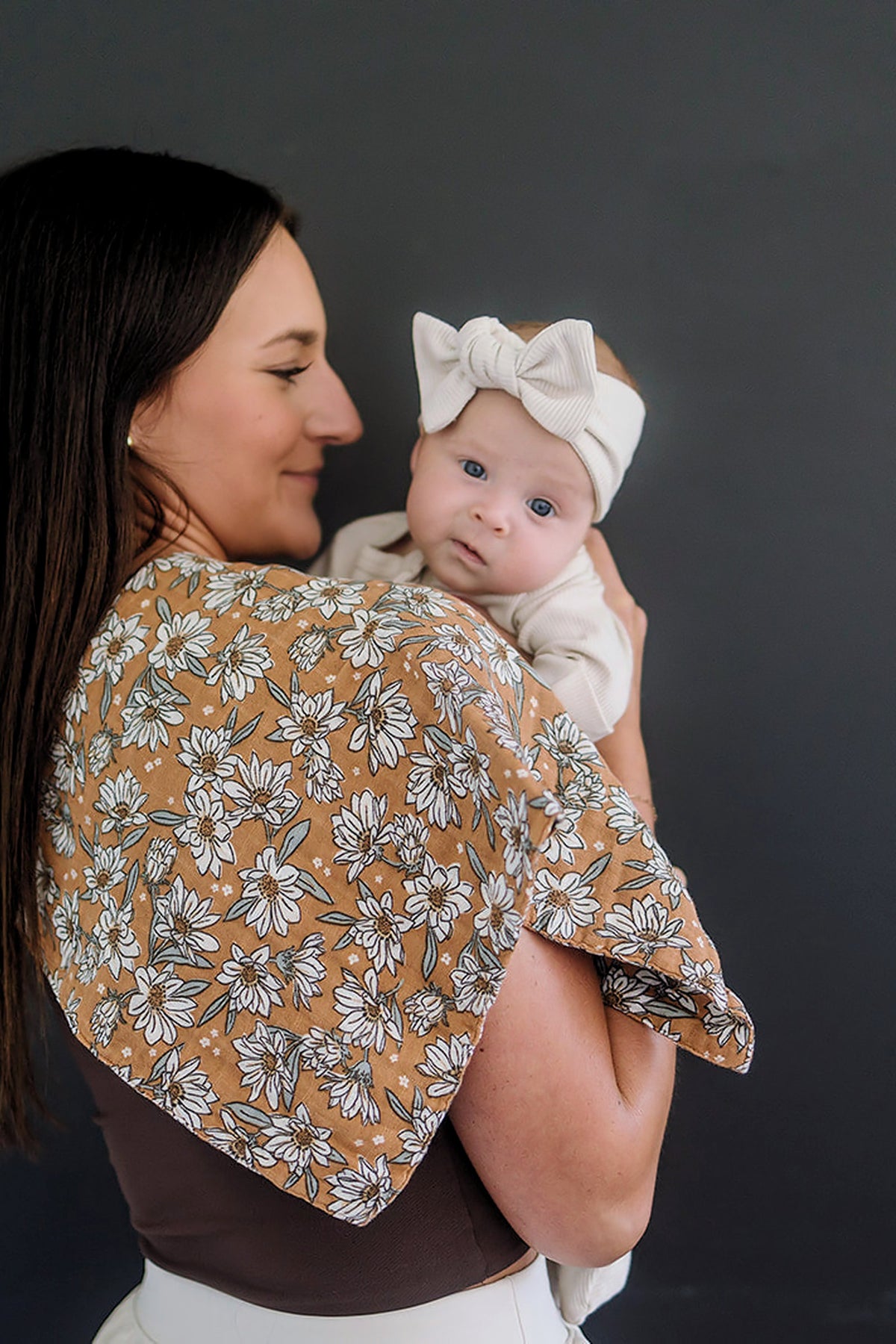 A woman holds a baby on her shoulder, using a floral muslin burp cloth. The baby wears a headband.