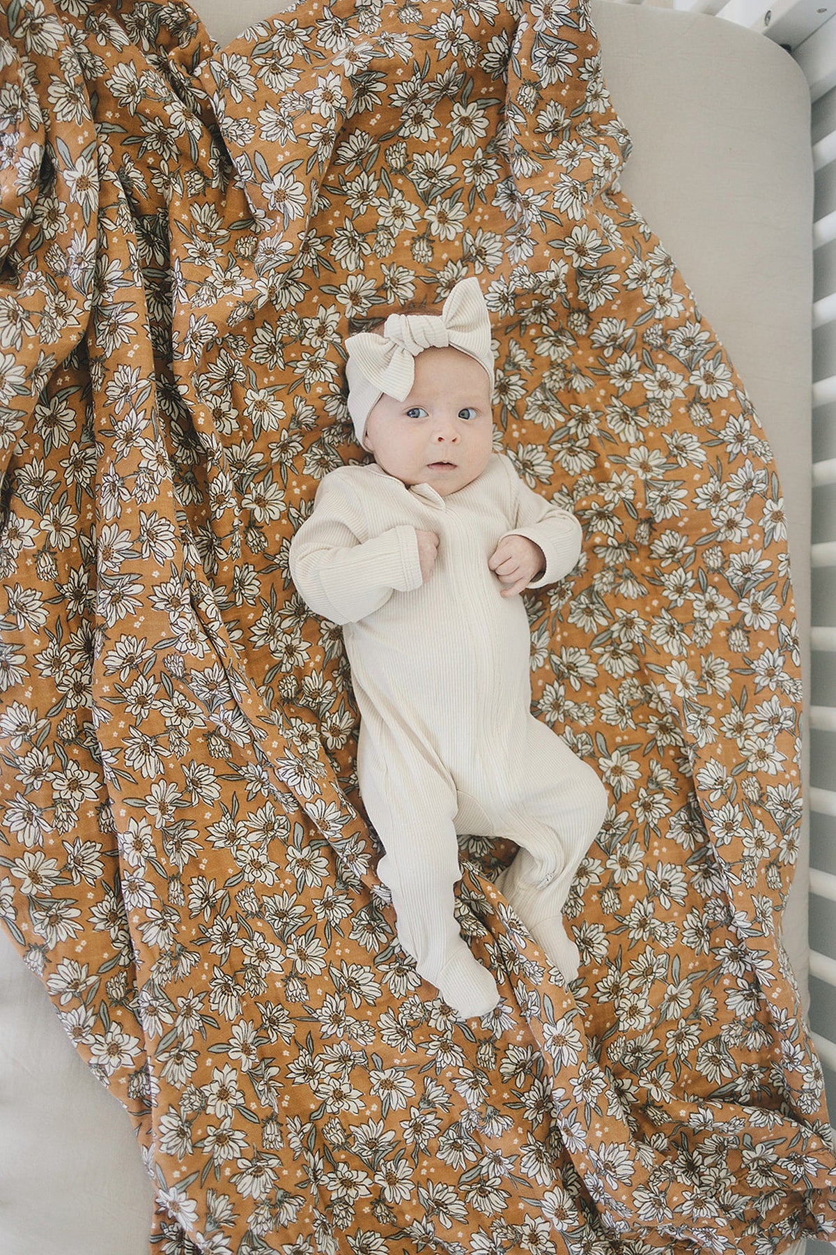 A baby in a cream outfit lies on a floral magnolia print muslin quilt in a crib.