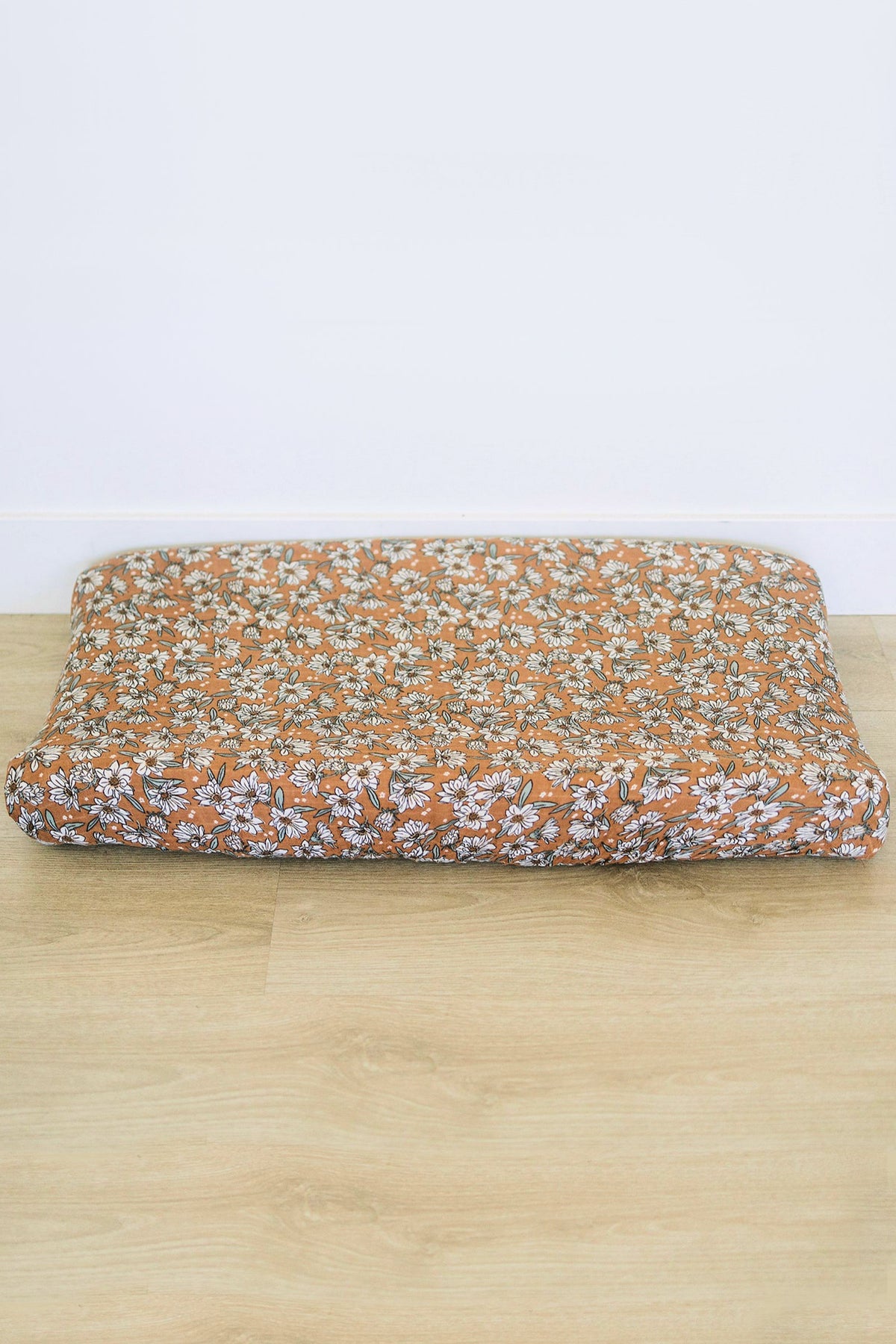 Floral-patterned muslin changing pad cover with white flowers on a brown background, placed on a wooden floor.