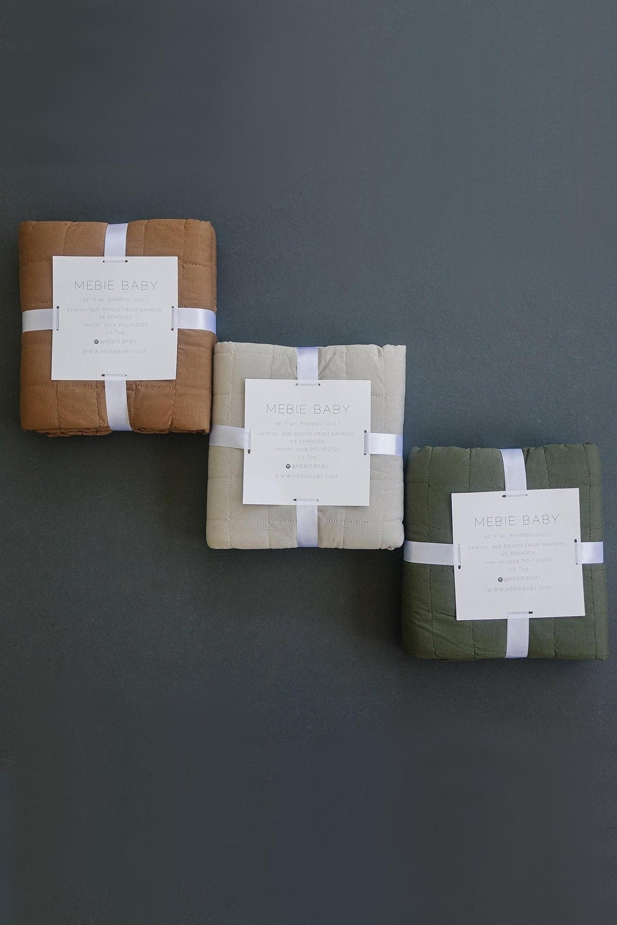 Three folded Mebie Baby bamboo quilts in brown, beige, and olive, tied with white ribbons on a dark surface.