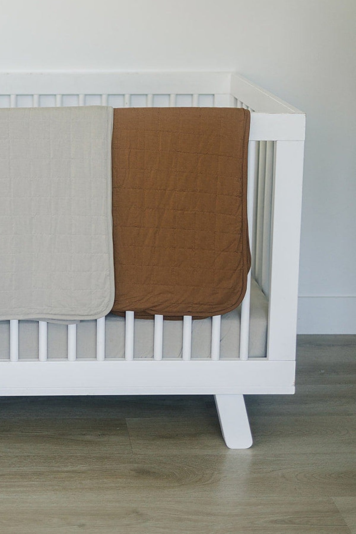 Two quilts, one beige and one mustard, neatly draped over a white crib in a bright room.