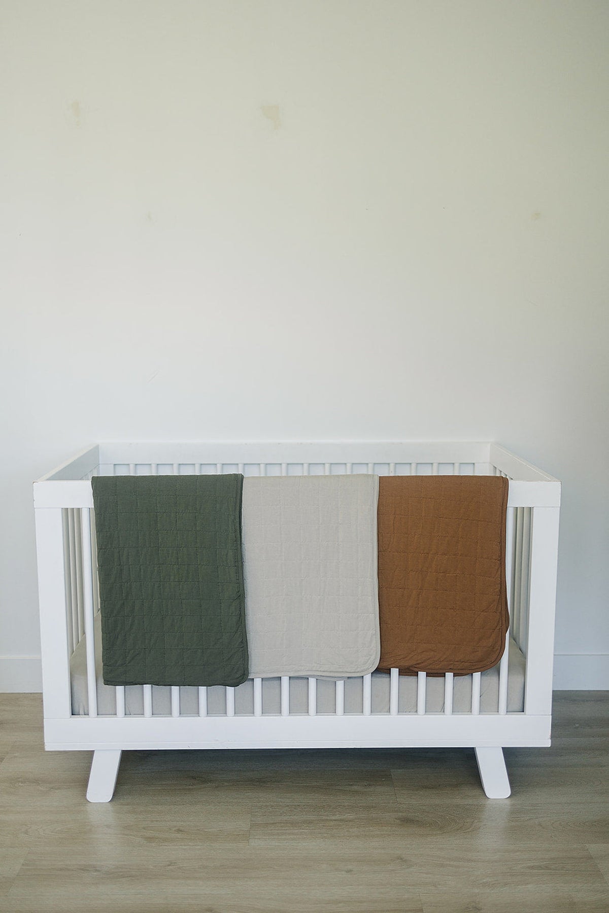 Three quilts in green, oatmeal, and brown draped over a white crib, set against a plain wall.