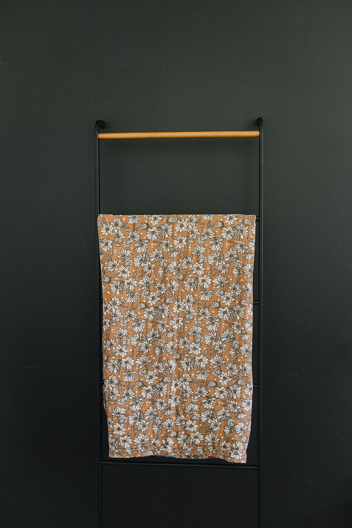 Floral-patterned brown quilt hangs on a black metal and wood ladder against a dark wall.