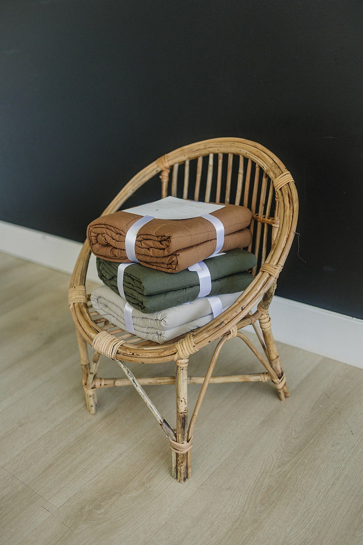 Three folded quilts in brown, green, and beige sit on a wicker chair against a black wall.