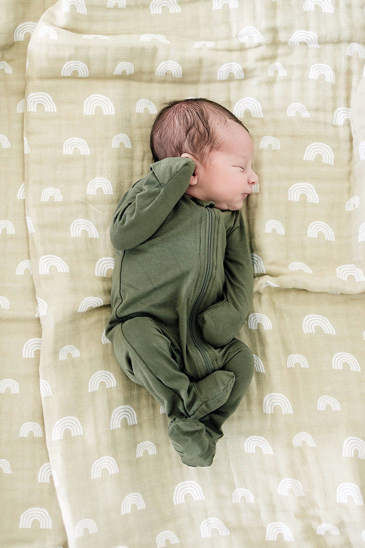 A baby sleeps peacefully in an olive green footed pajama on a patterned blanket with white rainbows.