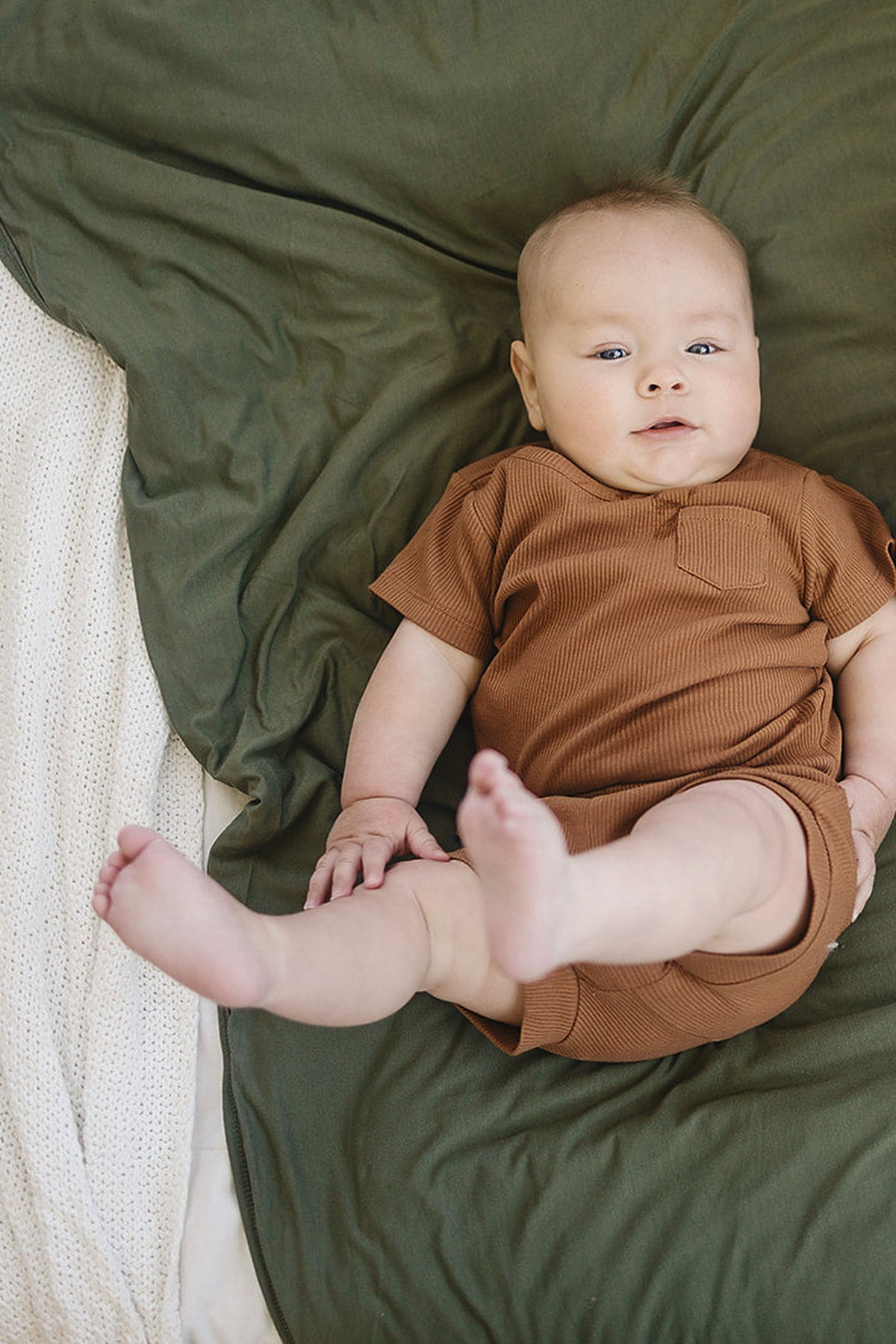 A baby in a brown outfit lies on an olive green quilt, with a white textured blanket nearby.