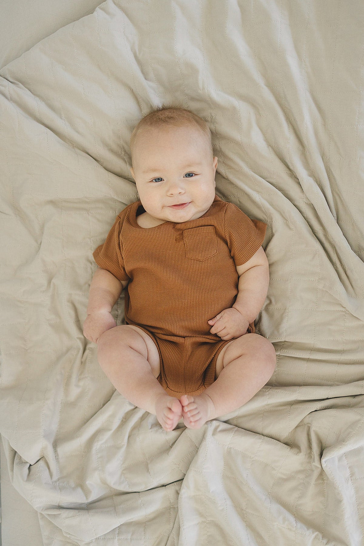 A baby in a brown onesie lies on a quilted oatmeal-colored blanket, smiling softly.