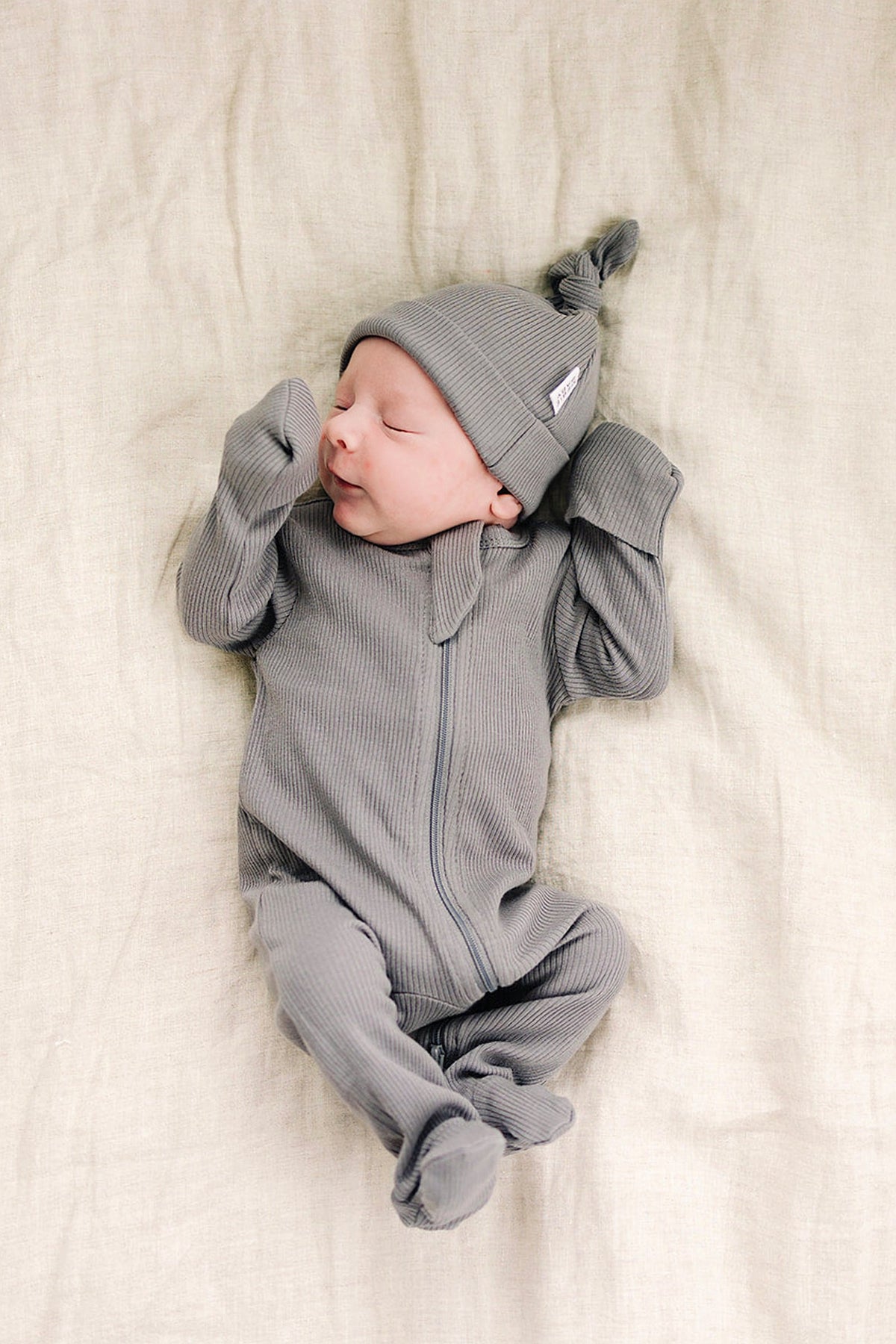 Newborn in grey ribbed footed pajama and hat, lying on a soft beige surface, featuring a zipper closure.