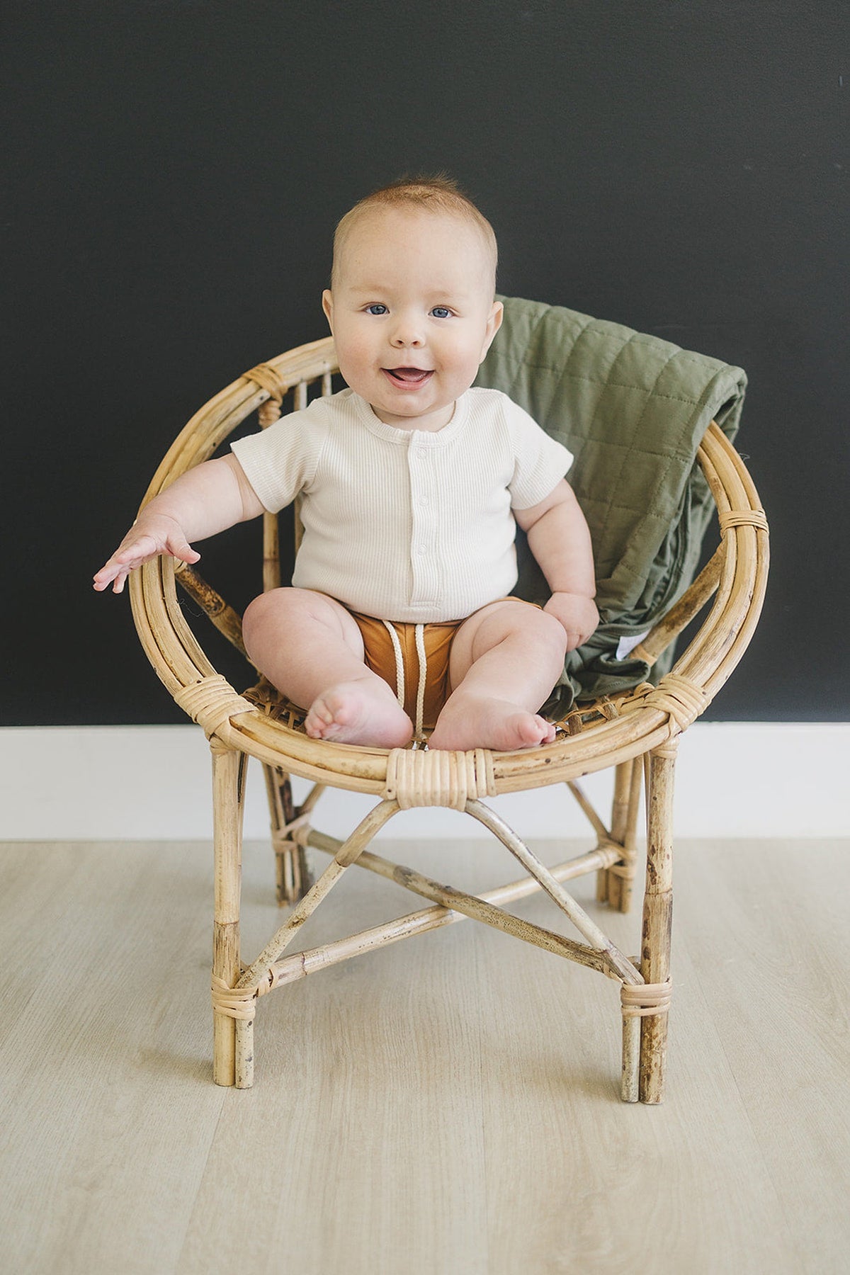 A baby in a white ribbed bodysuit sits on a bamboo chair with a green blanket draped over it.