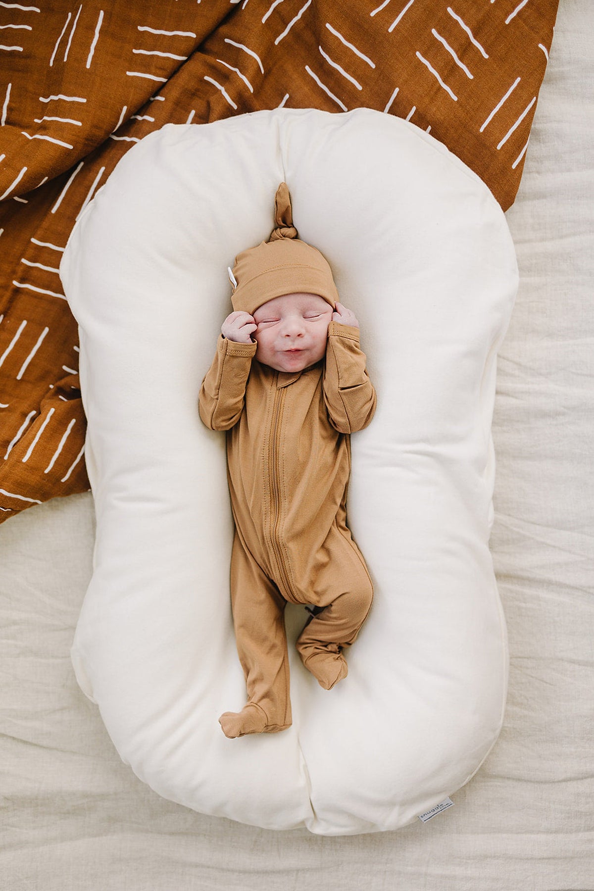 Newborn in a mustard bamboo knot hat and matching onesie, resting on a white cushion with a brown patterned blanket.