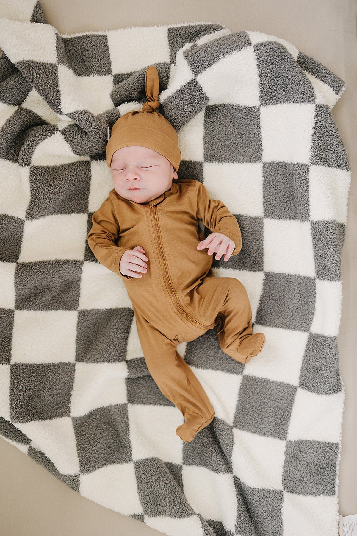 A baby wearing mustard footed pajamas and a matching hat lies on a black and white checkered blanket.