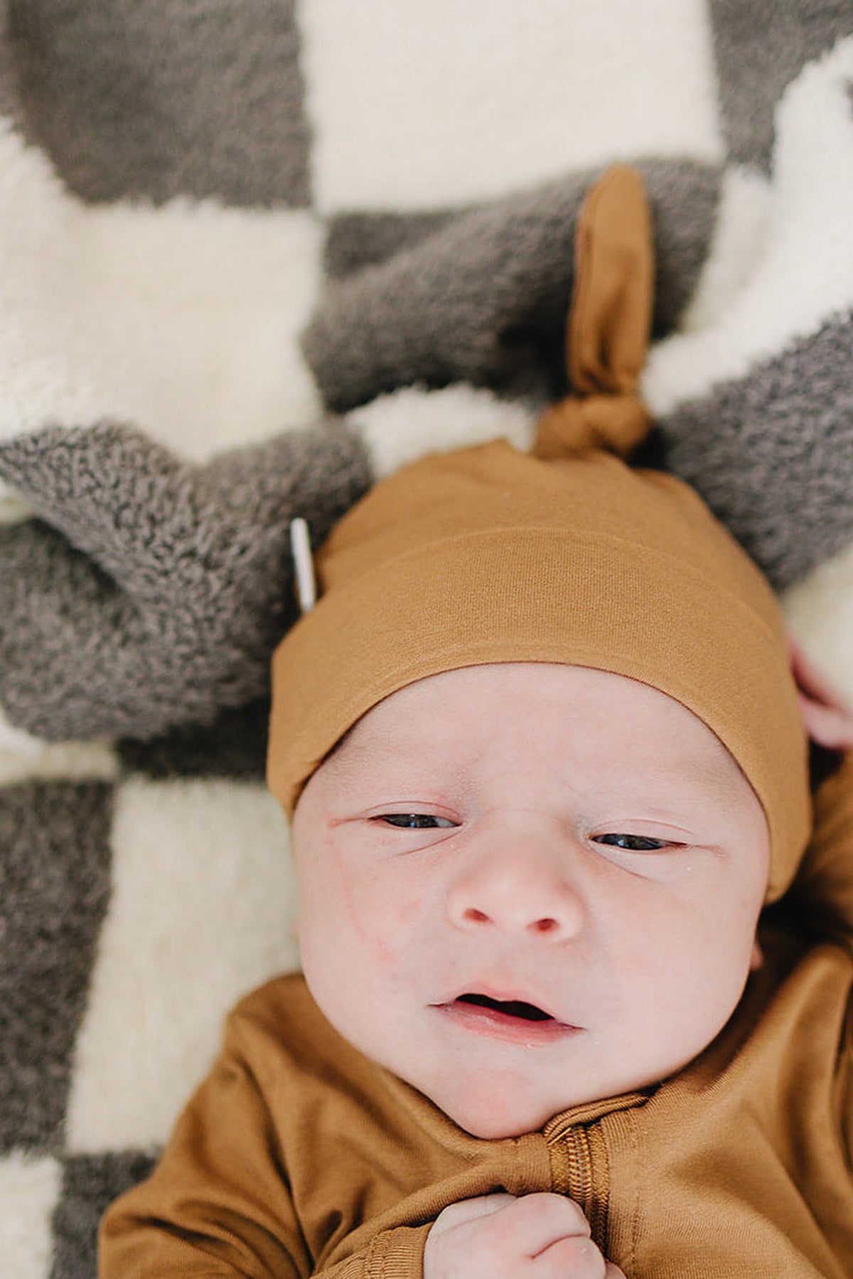 A newborn wearing a mustard knot hat lies on a soft checkered blanket, looking content and cozy.