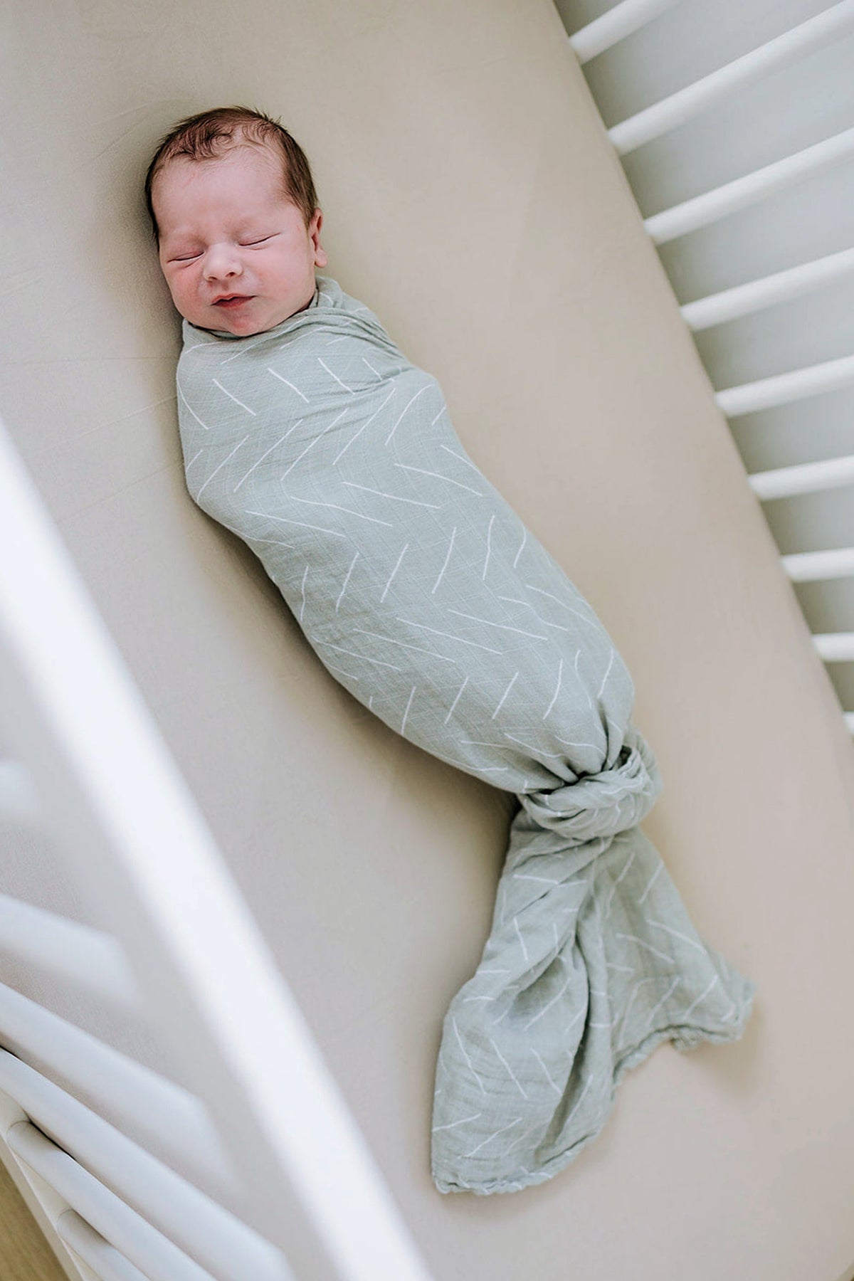 A baby is peacefully swaddled in a light green blanket with white patterns, lying in a crib.