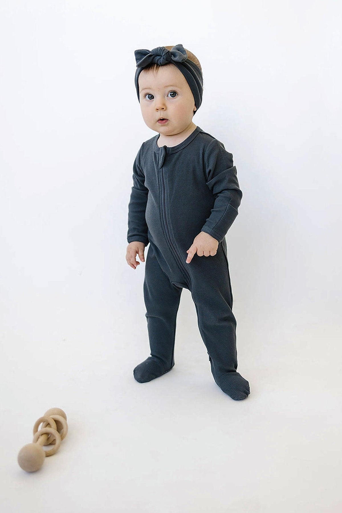 A baby in charcoal ribbed footed pajamas and headband stands beside wooden toys on a white background.
