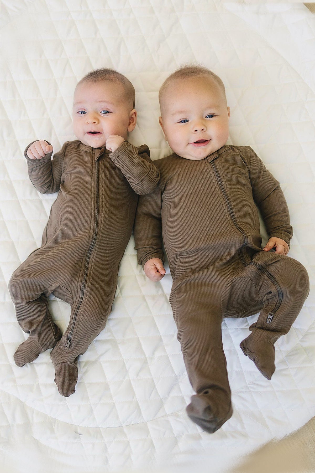 Two babies wearing cocoa-colored ribbed footed pajamas with zippers, lying on a white quilted surface.