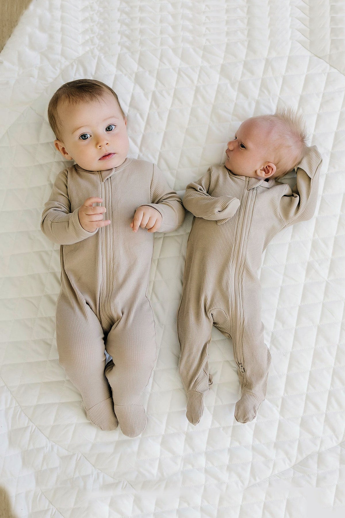 Two babies lying on a white quilt, wearing oatmeal-colored ribbed zipper footed pajamas made from organic cotton.