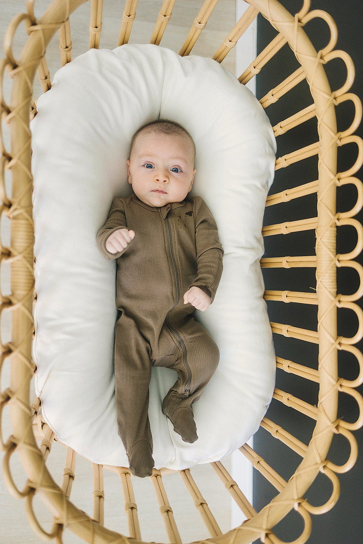 A baby in cocoa ribbed zipper pajamas lies in a wicker bassinet with a white cushion.