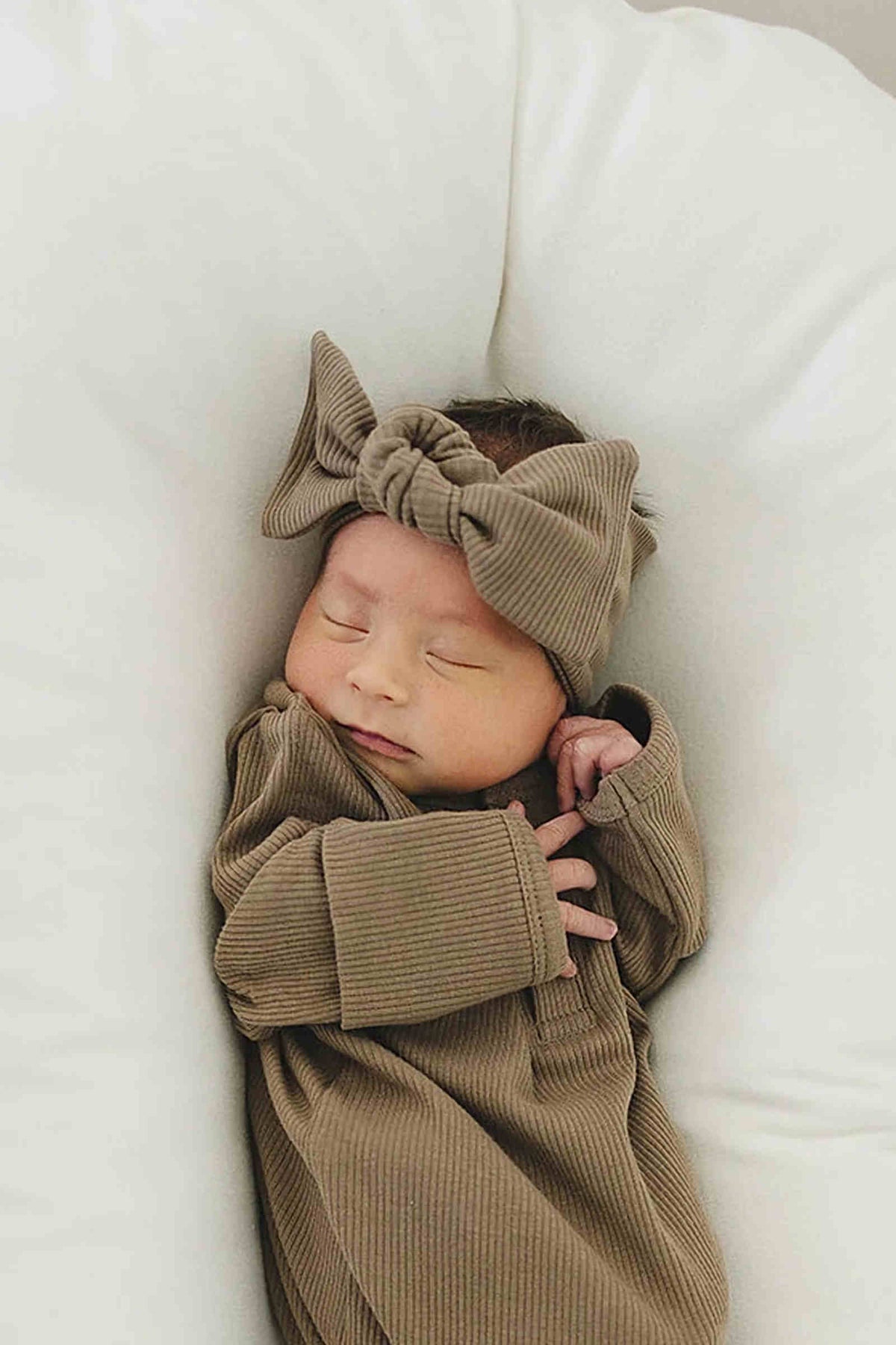 A sleeping baby wears a cocoa ribbed head wrap and matching outfit, nestled in a soft white cushion.