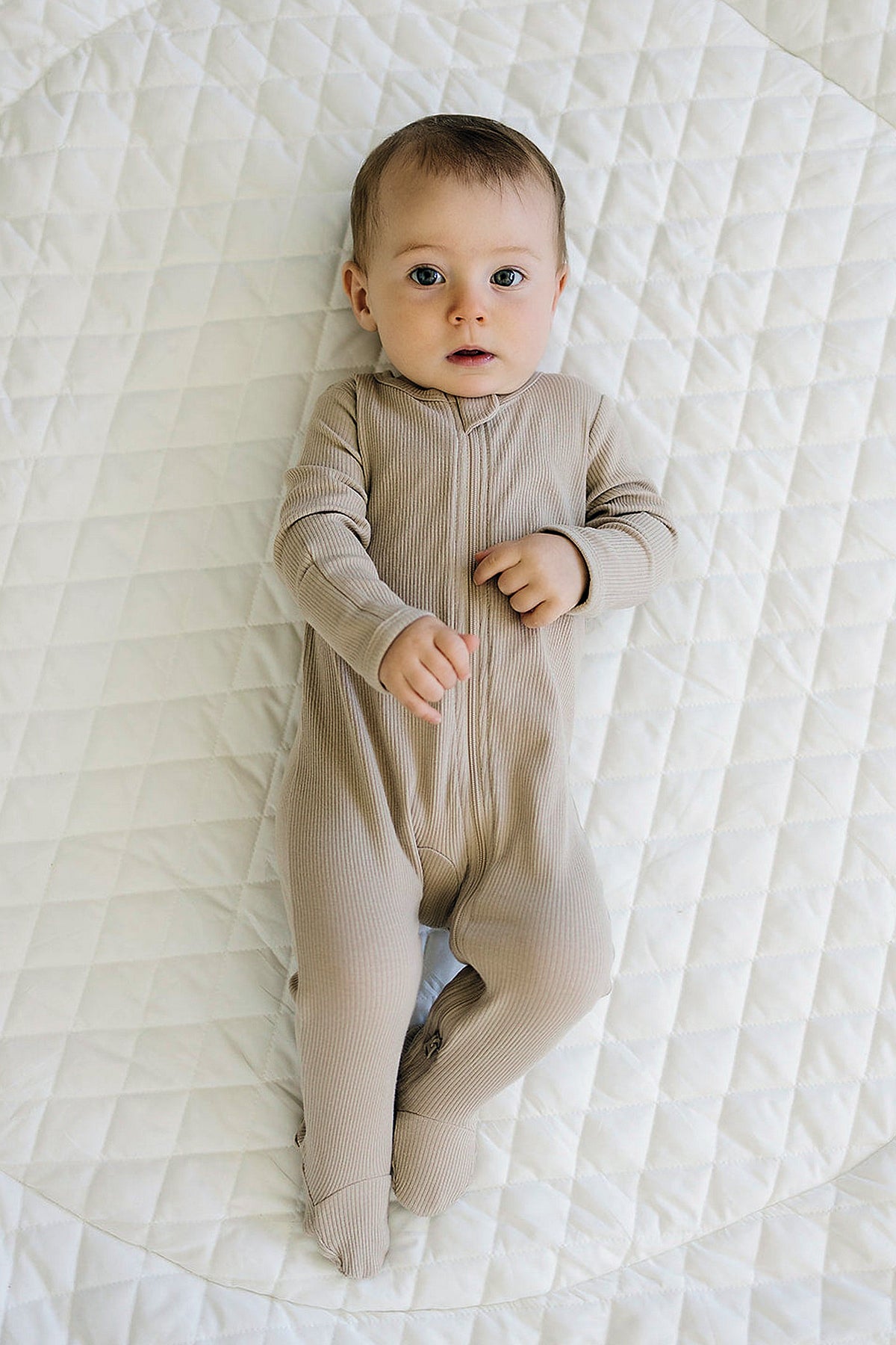 A baby wearing oatmeal-colored ribbed pajamas lies on a quilted white blanket, looking up.