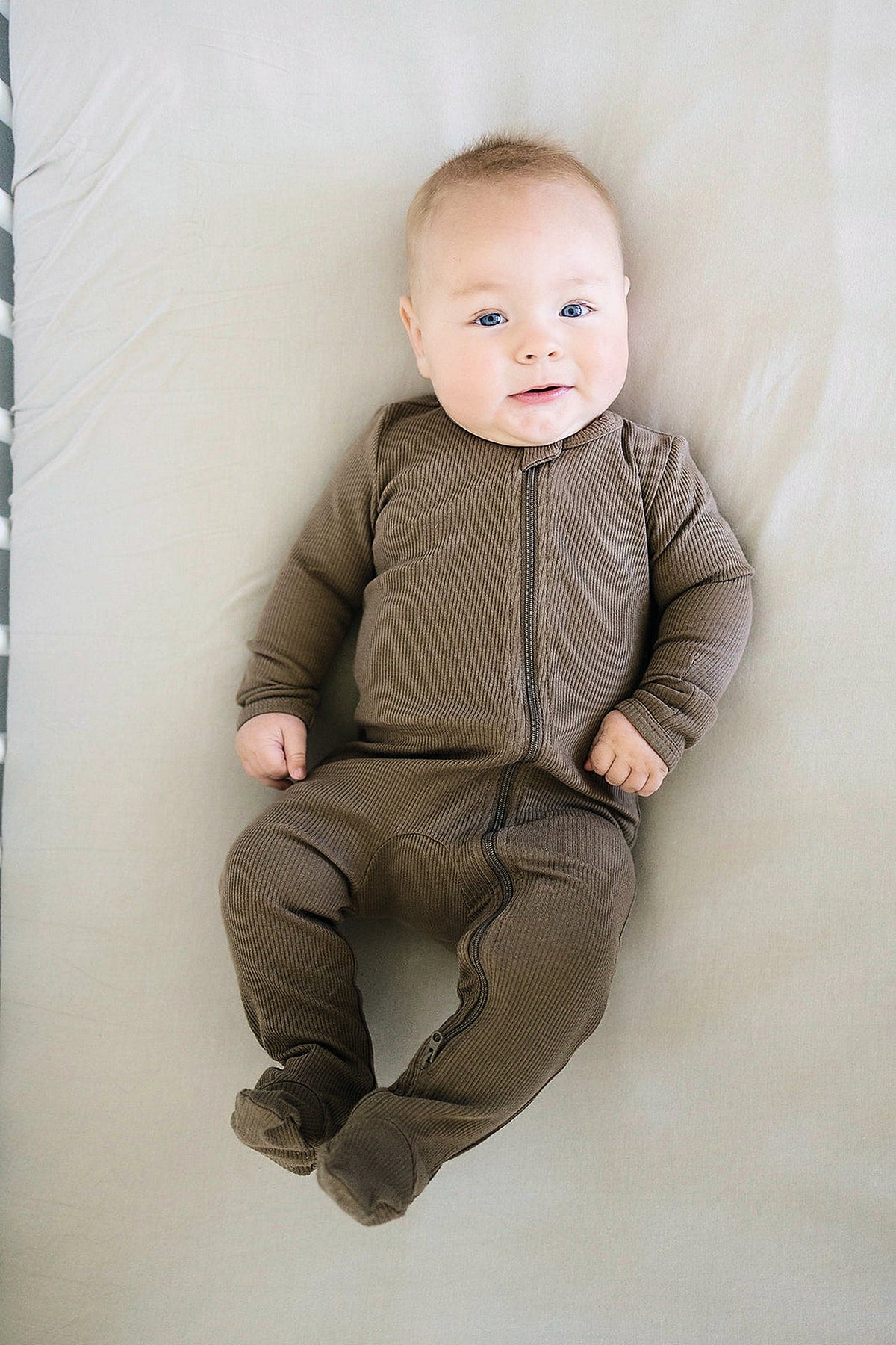 A baby wearing cocoa-colored, ribbed, footed pajamas with a zipper lies on a light-colored surface, smiling contentedly.
