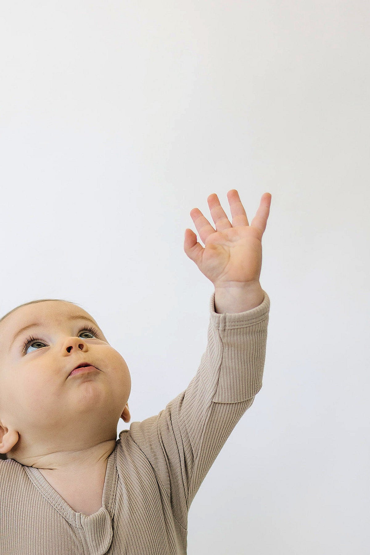 A baby in an oatmeal-colored ribbed pajama reaches upward against a plain white background.