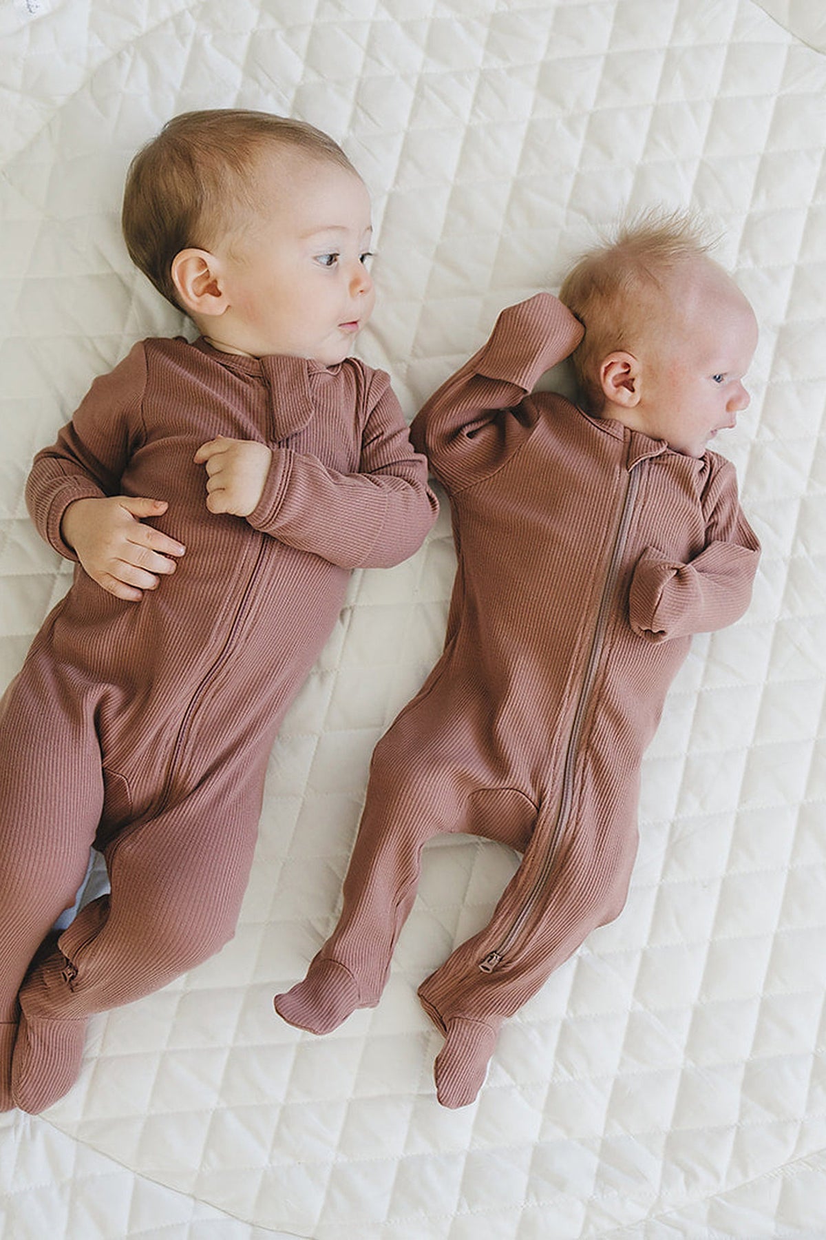 Two babies wearing dusty rose ribbed zipper footed pajamas lie on a white quilted surface.