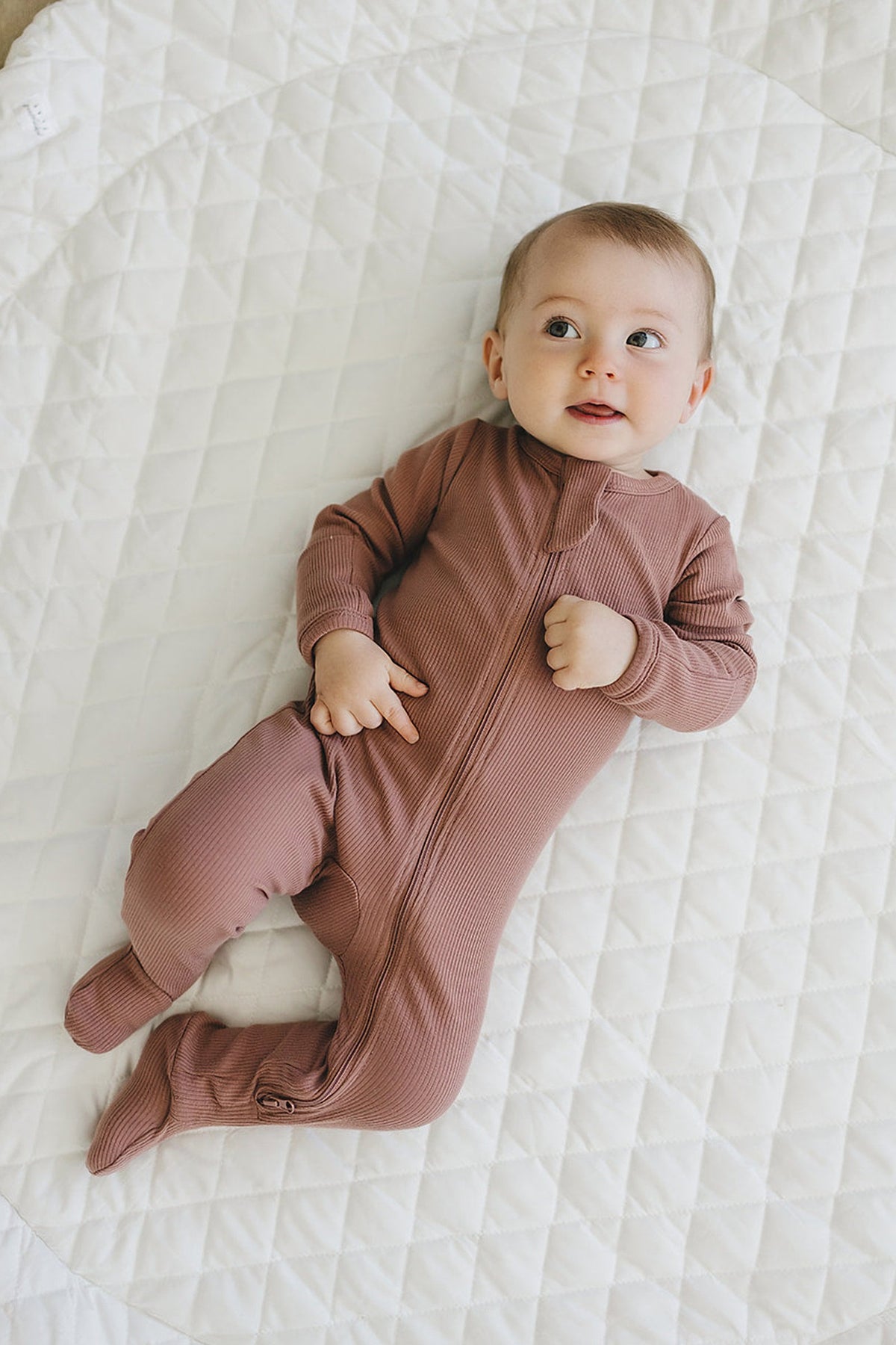 Baby in dusty rose ribbed zipper footed pajama lying on a white quilted blanket, looking upwards.