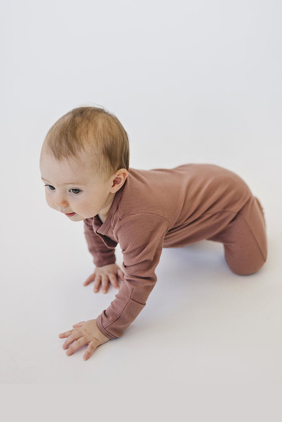 A baby in dusty rose ribbed pajamas crawls on a white background, showcasing the garments soft texture.