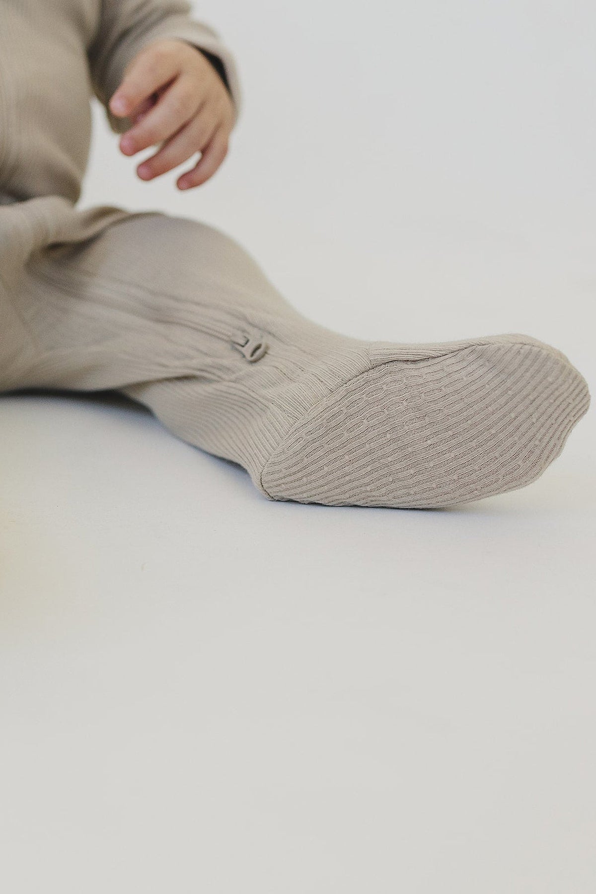 A baby wearing oatmeal-colored ribbed footed pajamas with a visible zipper, sitting on a light surface.
