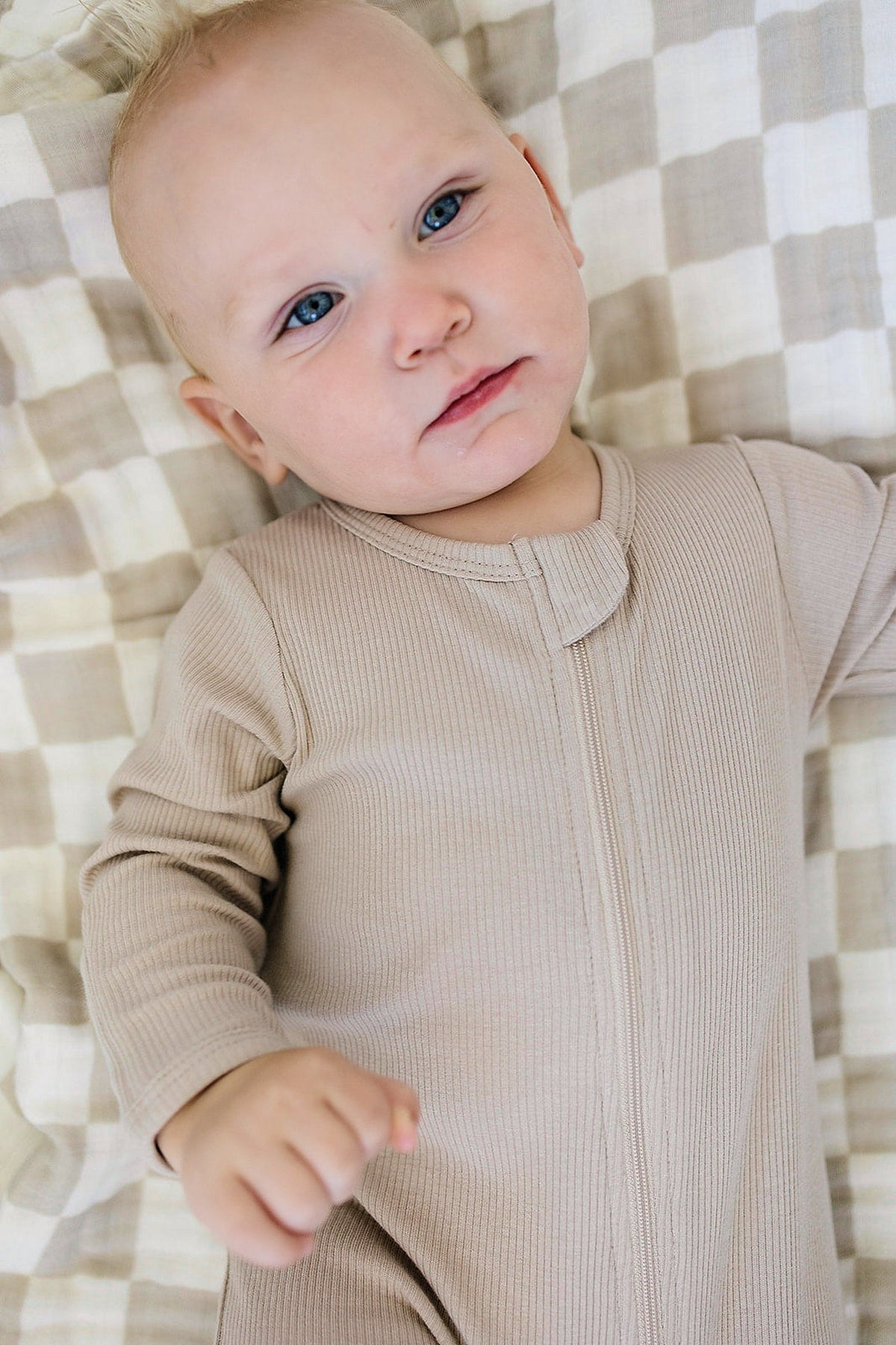 Baby in oatmeal ribbed zipper footed pajama lying on a checkered blanket, featuring organic cotton fabric.