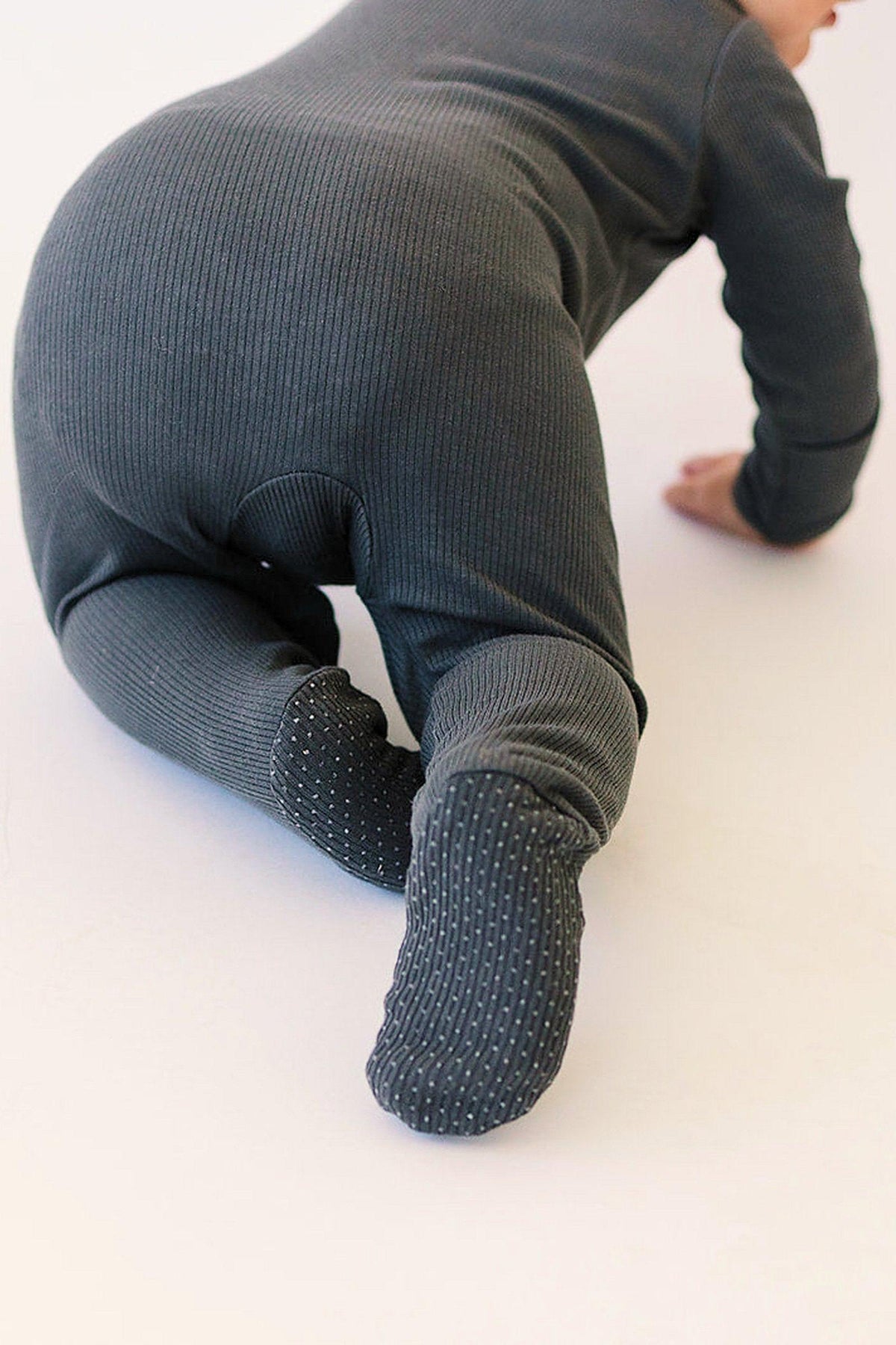 A baby in charcoal ribbed pajamas crawls on a light surface, showcasing the footed design with grip details.