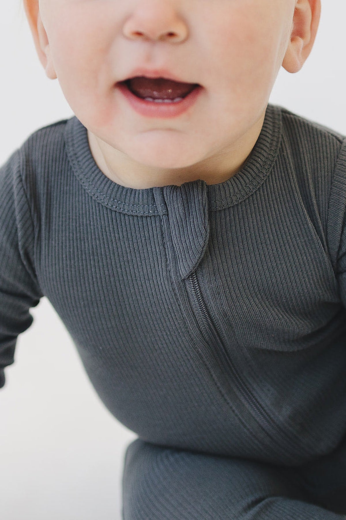 A baby wearing a ribbed, charcoal-colored zipper pajama made from organic cotton, showing a close-up of the garment.