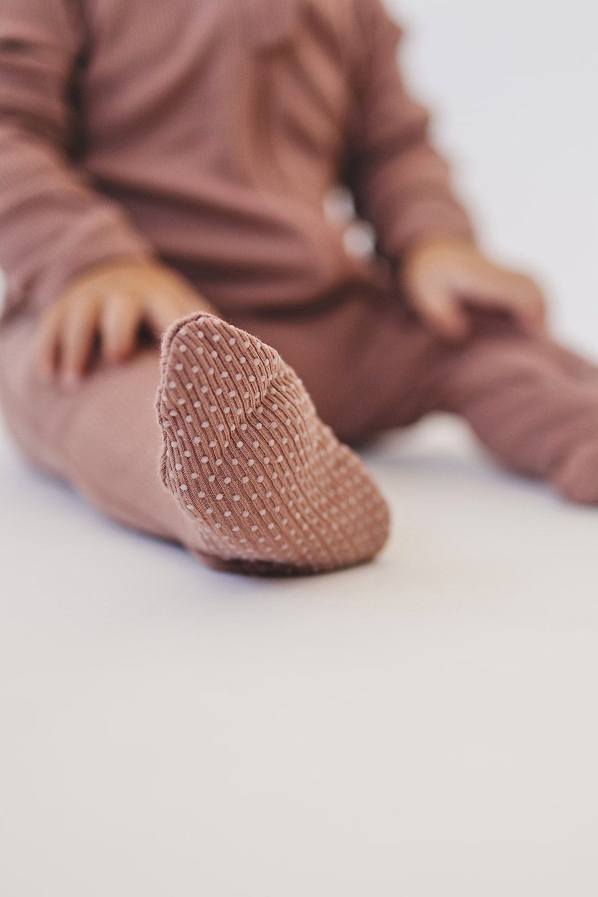 A baby wearing dusty rose ribbed pajamas with a footed design, featuring a textured non-slip sole.
