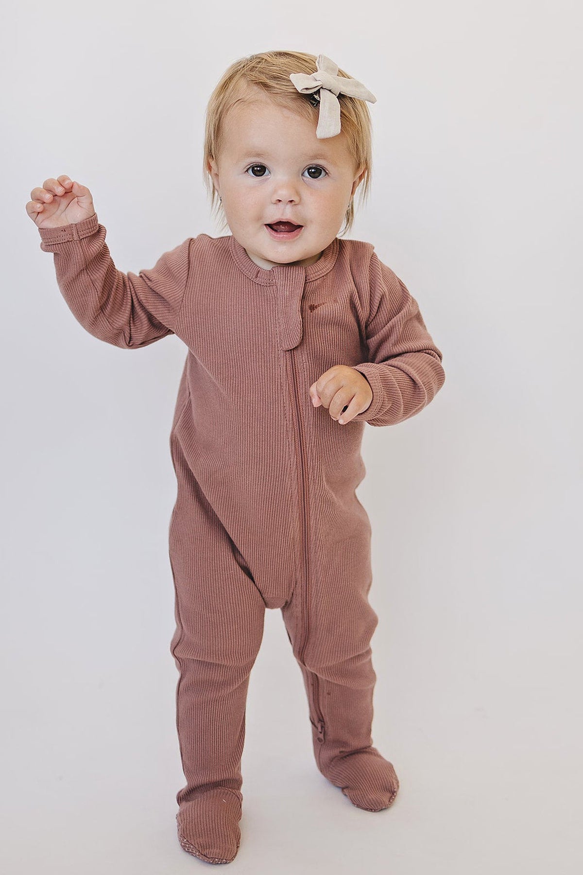 A baby wearing a dusty rose ribbed zipper footed pajama with a white bow on their head.