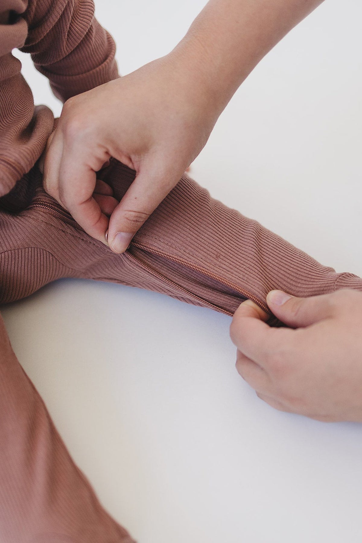 Hands zipping dusty rose ribbed baby pajamas made from organic cotton on a white background.