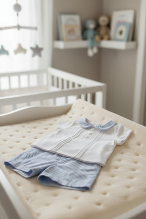 A white short-sleeve shirt with a blue-striped collar is paired with matching blue-striped shorts, displayed on a soft changing pad.